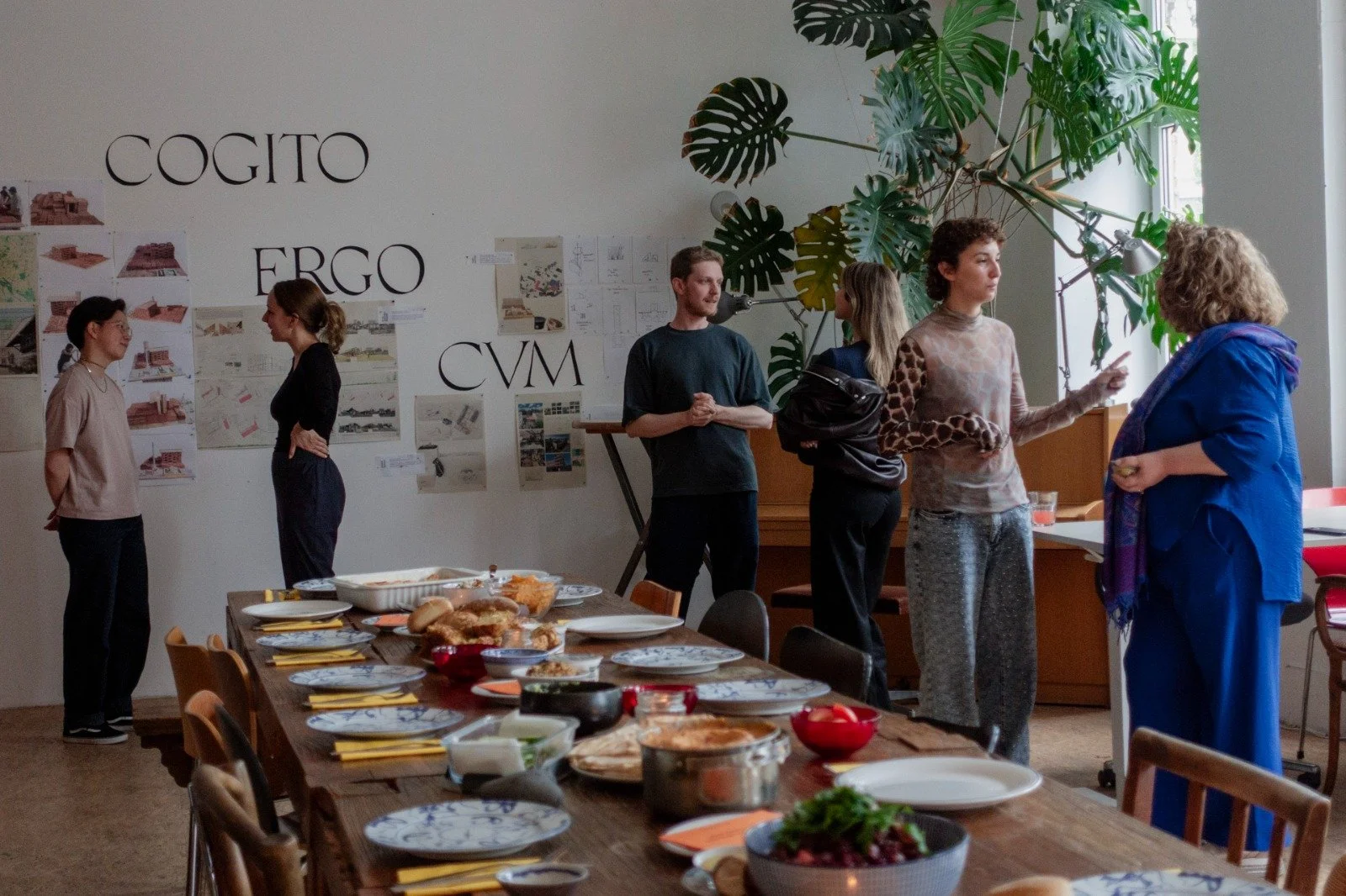 People standing and talking in a room with a large dining table set with various dishes, plates, and utensils. A large green plant is visible near the window, and the wall has large text that reads 'COGITO ERGO CVM' with images and notes pinned nearb