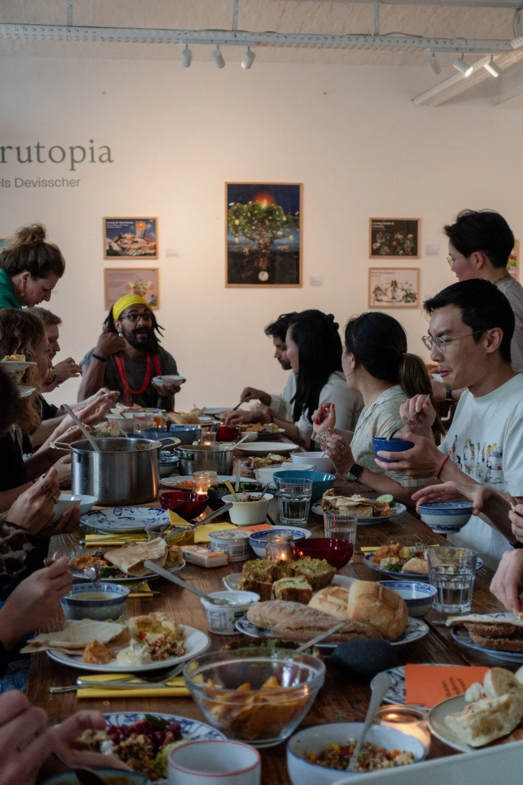 A diverse group of people gathered around a long table enjoying a meal together in an art gallery with framed artwork on the wall.