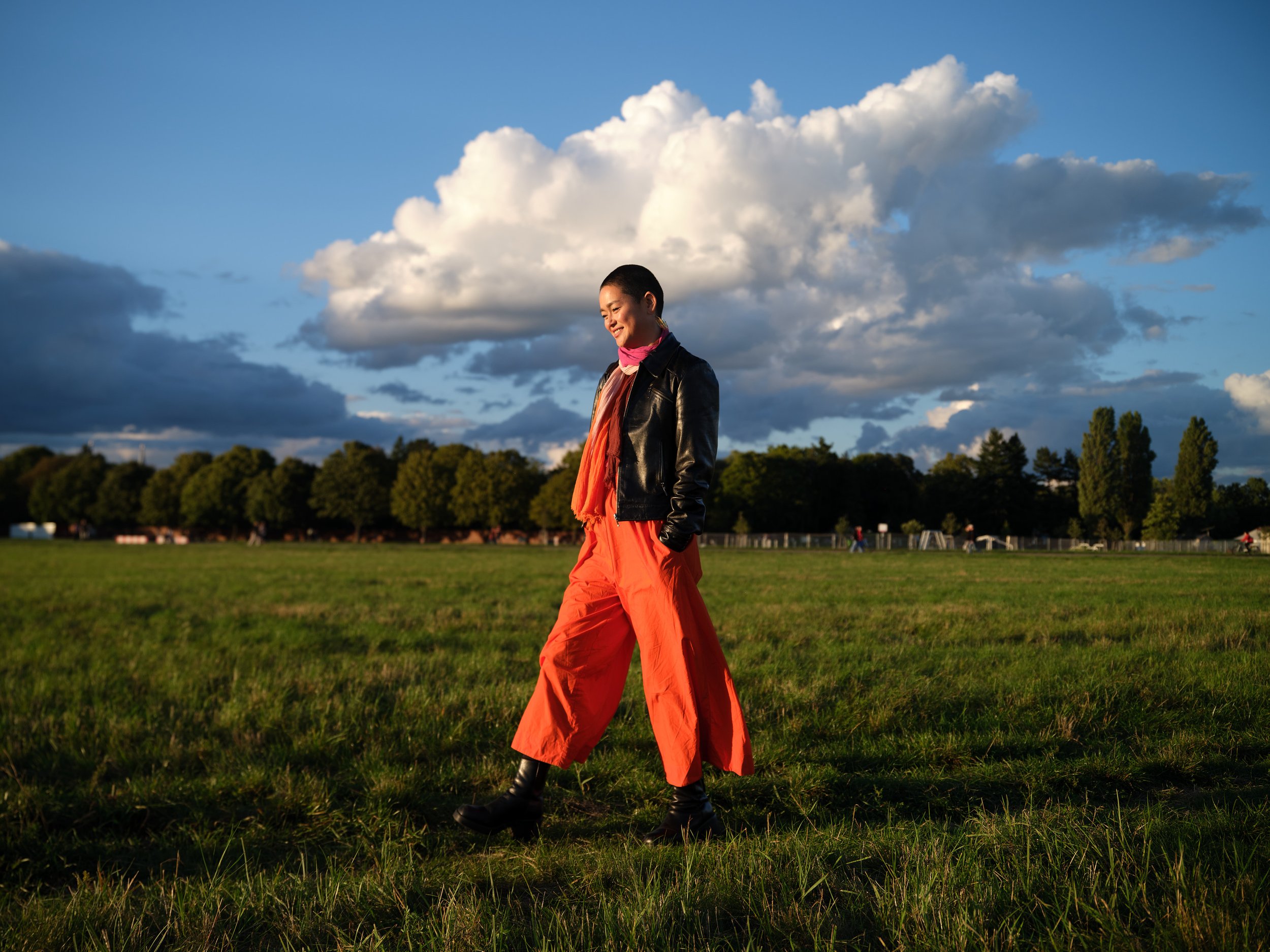 A woman with short hair wearing a black leather jacket, pink scarf, orange shirt, and bright orange pants standing outdoors on grass with a cloudy sky and distant trees in the background.