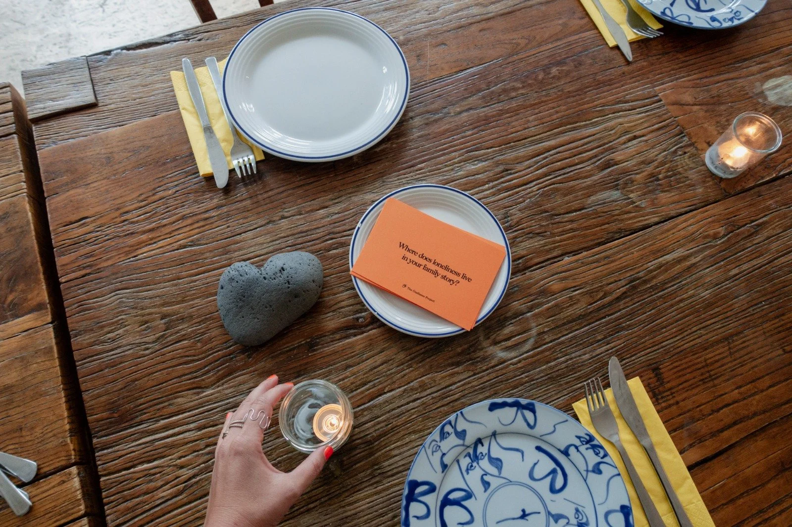 A wooden dining table set for a meal with blue and white plates, yellow napkins, and silverware. There are two small candles and a grey heart-shaped stone on the table. An orange card with the question, 'Where does loneliness live in your family story?' is on one plate.