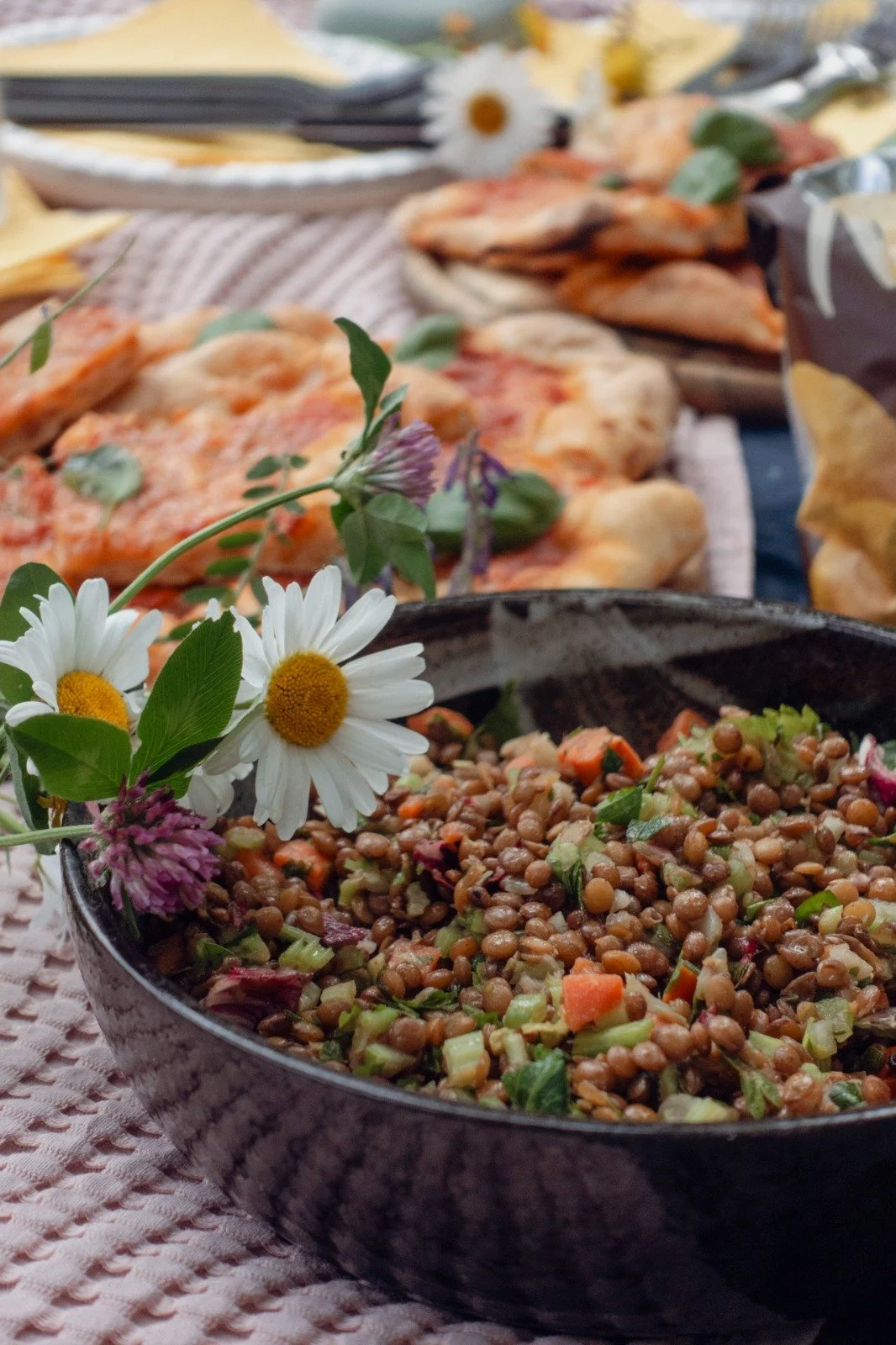 Bowl of lentil salad with chopped vegetables and herbs, topped with daisies and purple flowers, with pizza and chips in the background.