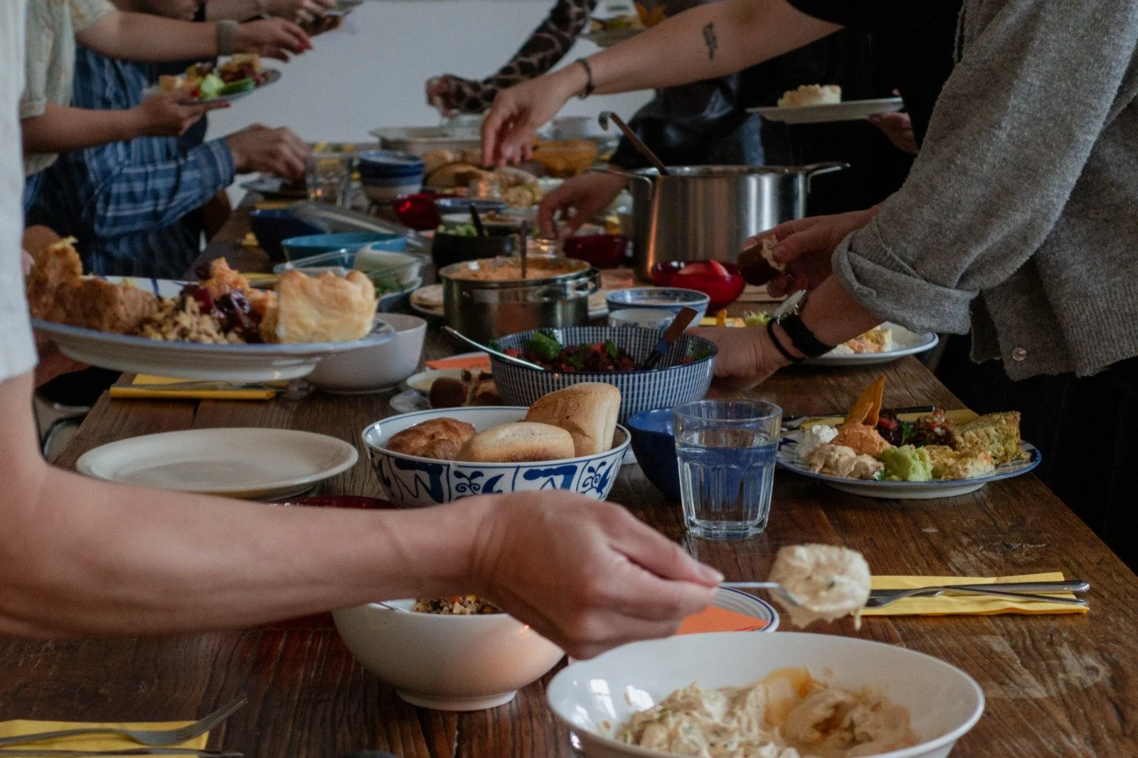 People gathered around a rustic wooden table enjoying a meal, with various dishes including bread, salads, and desserts, in a cozy indoor setting.