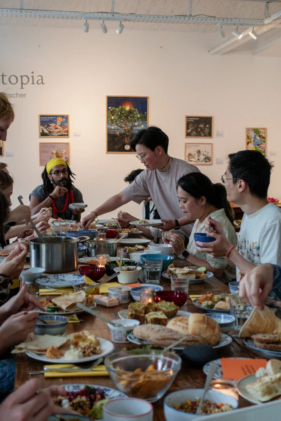 Group of diverse people at a long dining table, sharing a meal with various dishes, bowls, and drinks, in a well-lit art gallery or museum setting.