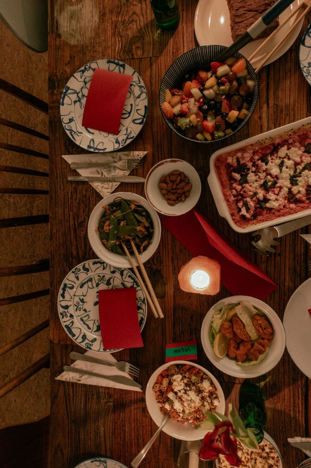 A table set for a meal with various dishes including a vegetable salad, a casserole dish with cheese and herbs, a bowl of almonds, a plate of fried chicken wings with lemon wedges, and a bowl of minced meat with cheese. There are plates, utensils, re