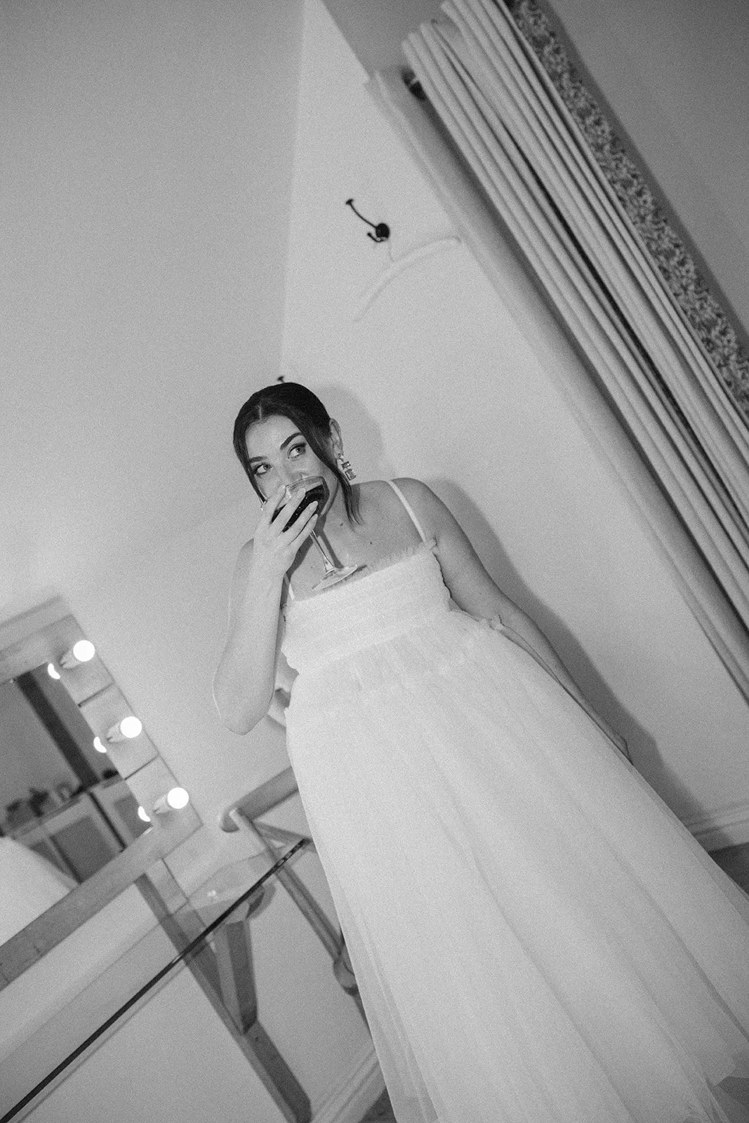 A woman in a white dress drinking from a wine glass in a room with mirror and vanity lights.