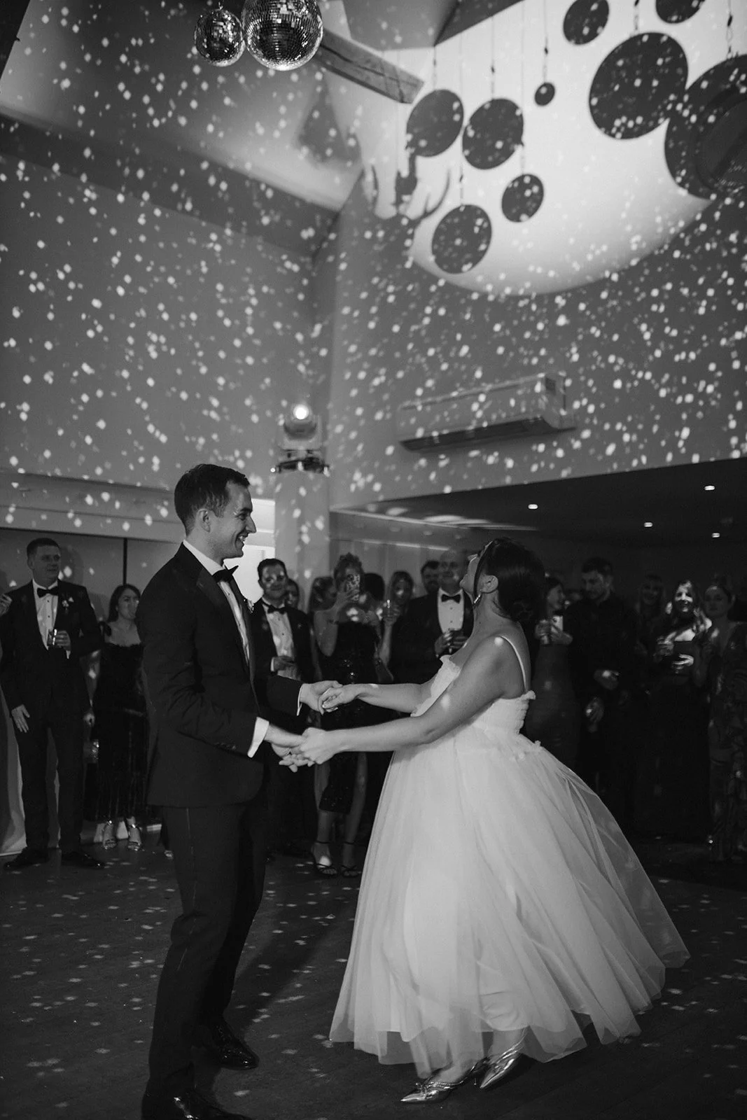 A black and white photo of a bride and groom dancing at their wedding reception surrounded by guests, with disco balls and hanging decorations overhead.