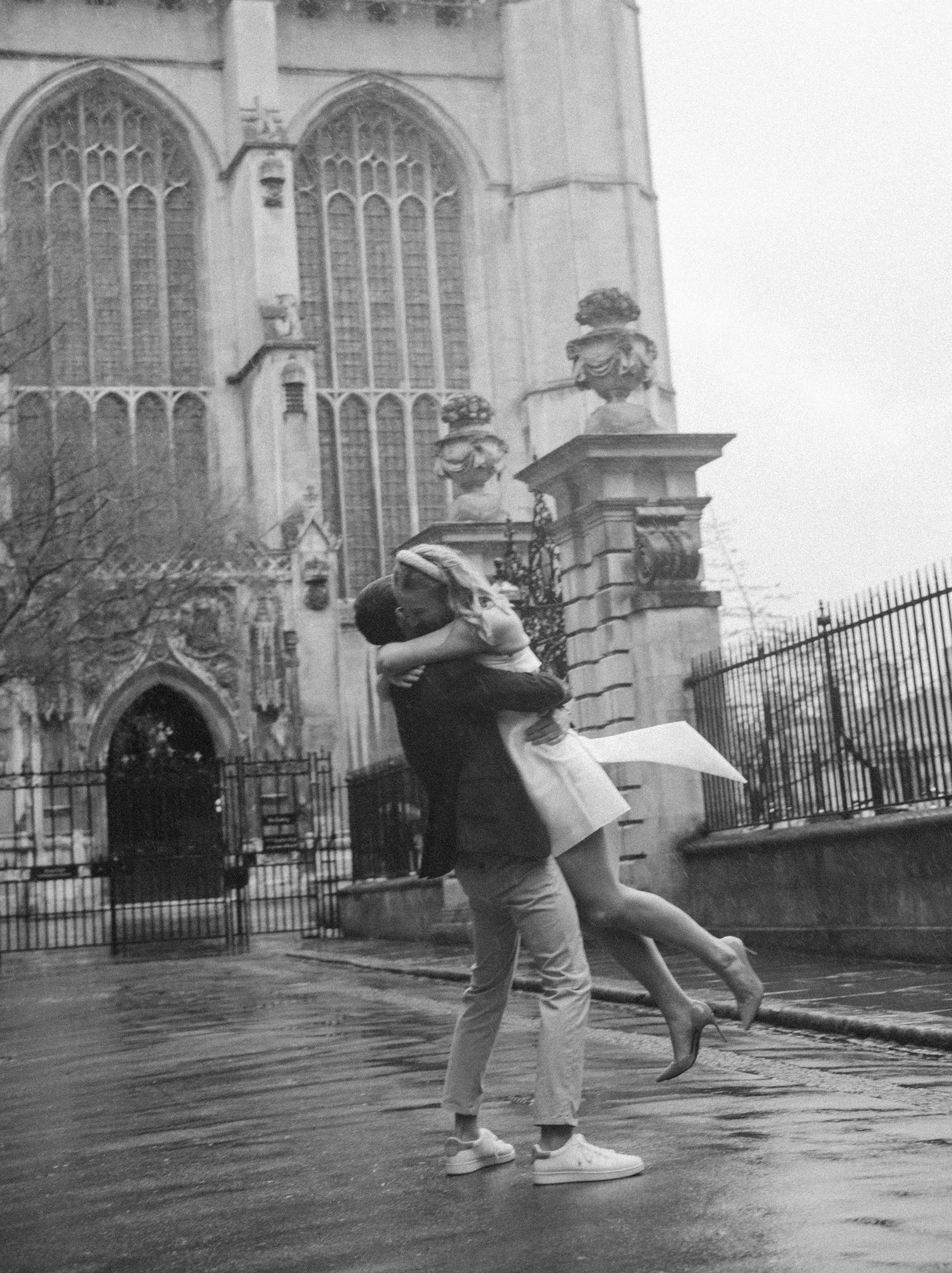 A man lifting a woman in front of a historic church. The woman is wearing high heels and a light-colored dress, and the man is wearing casual clothes. The scene appears romantic, and they are embracing as they stand on a wet street.