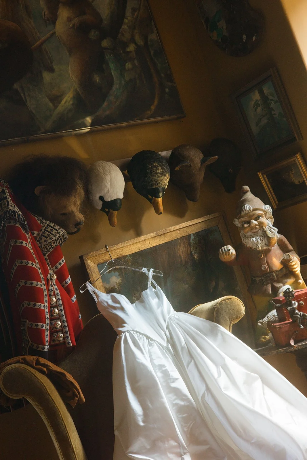 A white dress laid on an armchair in a room with various decorative items, including animal head masks on the wall, a Santa Claus figurine, framed paintings, and a red patterned jacket hanging on the armrest.
