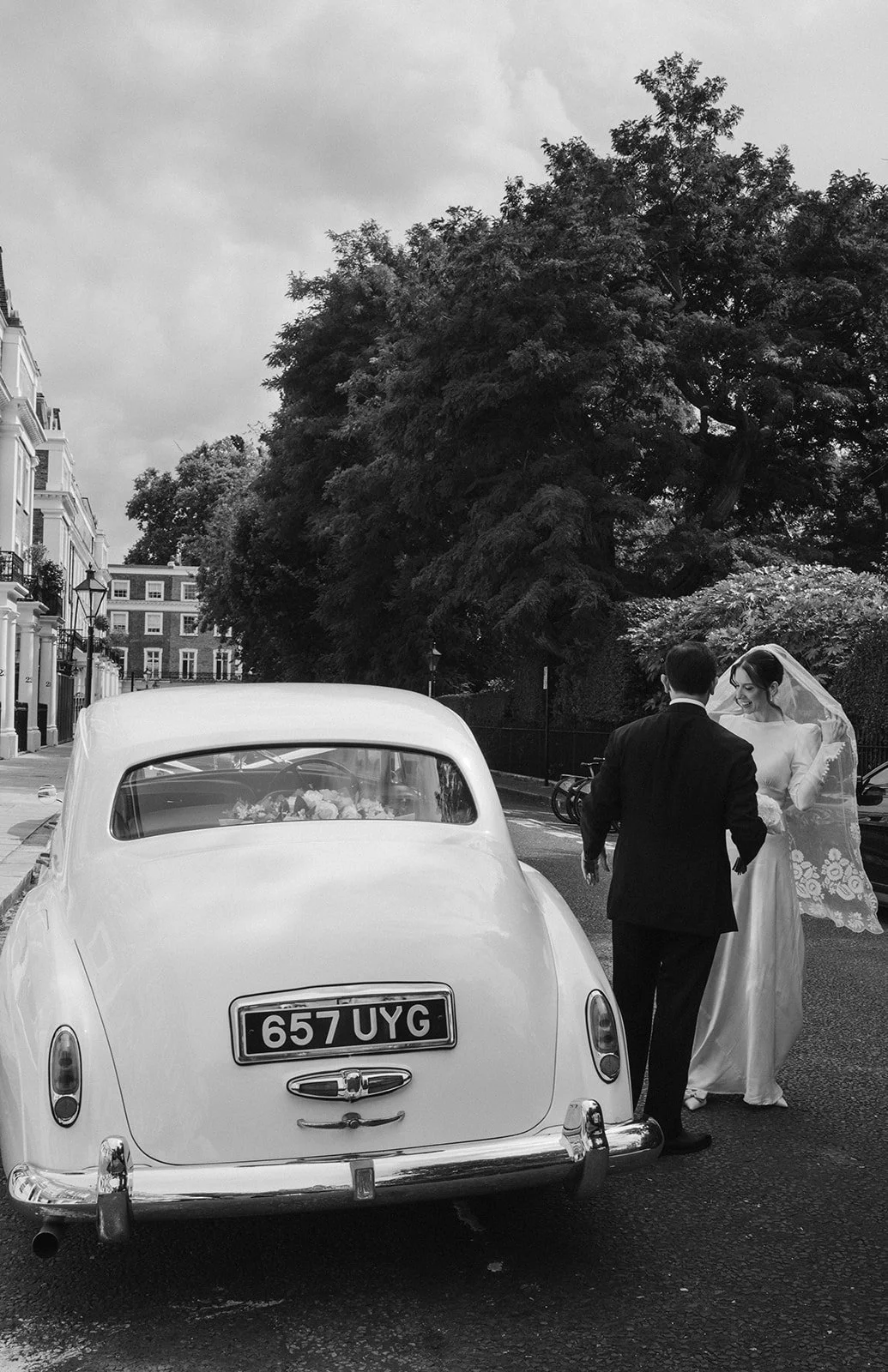A black and white photo of a bride and groom standing next to a vintage car on a city street, with large trees and buildings in the background.
