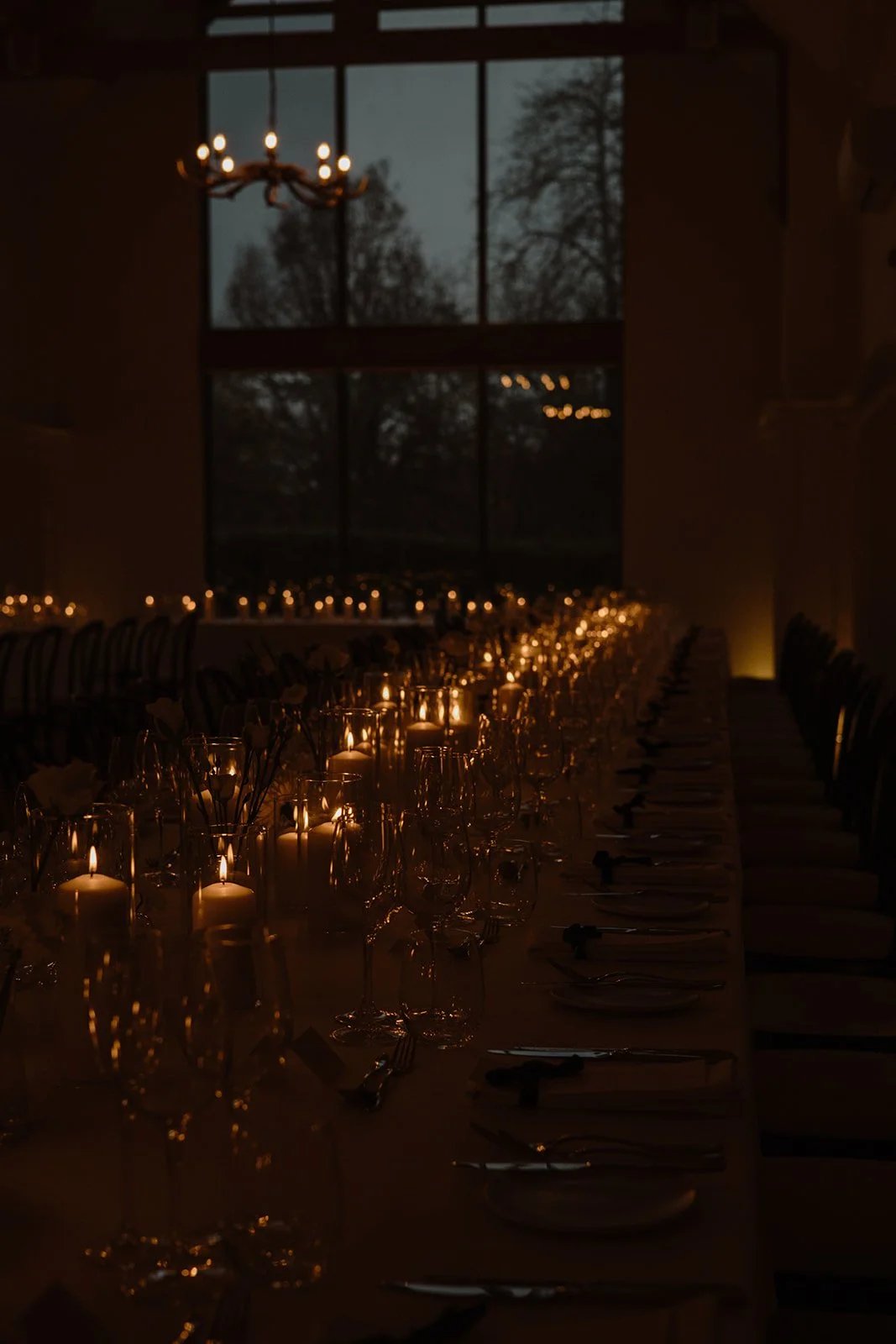 A dimly lit indoor dining area with a long table set for a formal dinner, adorned with candles, wine glasses, plates, and silverware, with large windows revealing a dark, cloudy sky and trees outside.