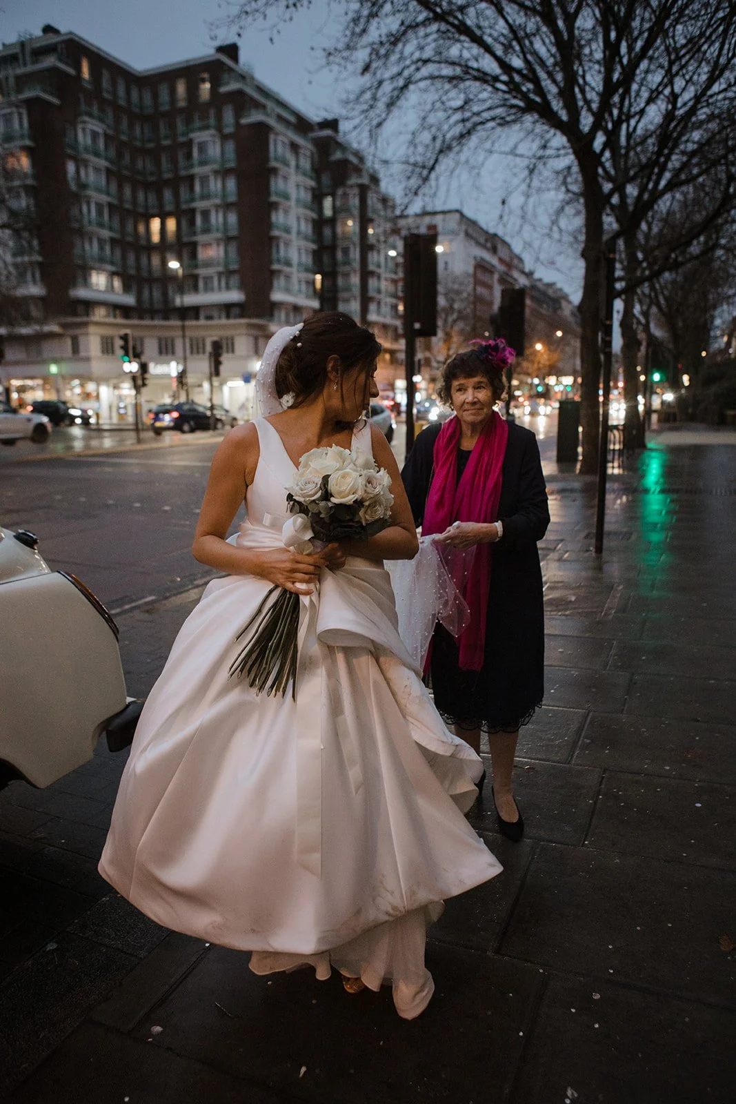 A bride holding a bouquet of white roses stands on a city sidewalk at dusk, with an older woman dressed in black with a pink scarf and matching headpiece next to her. Buildings and streetlights are visible in the background.