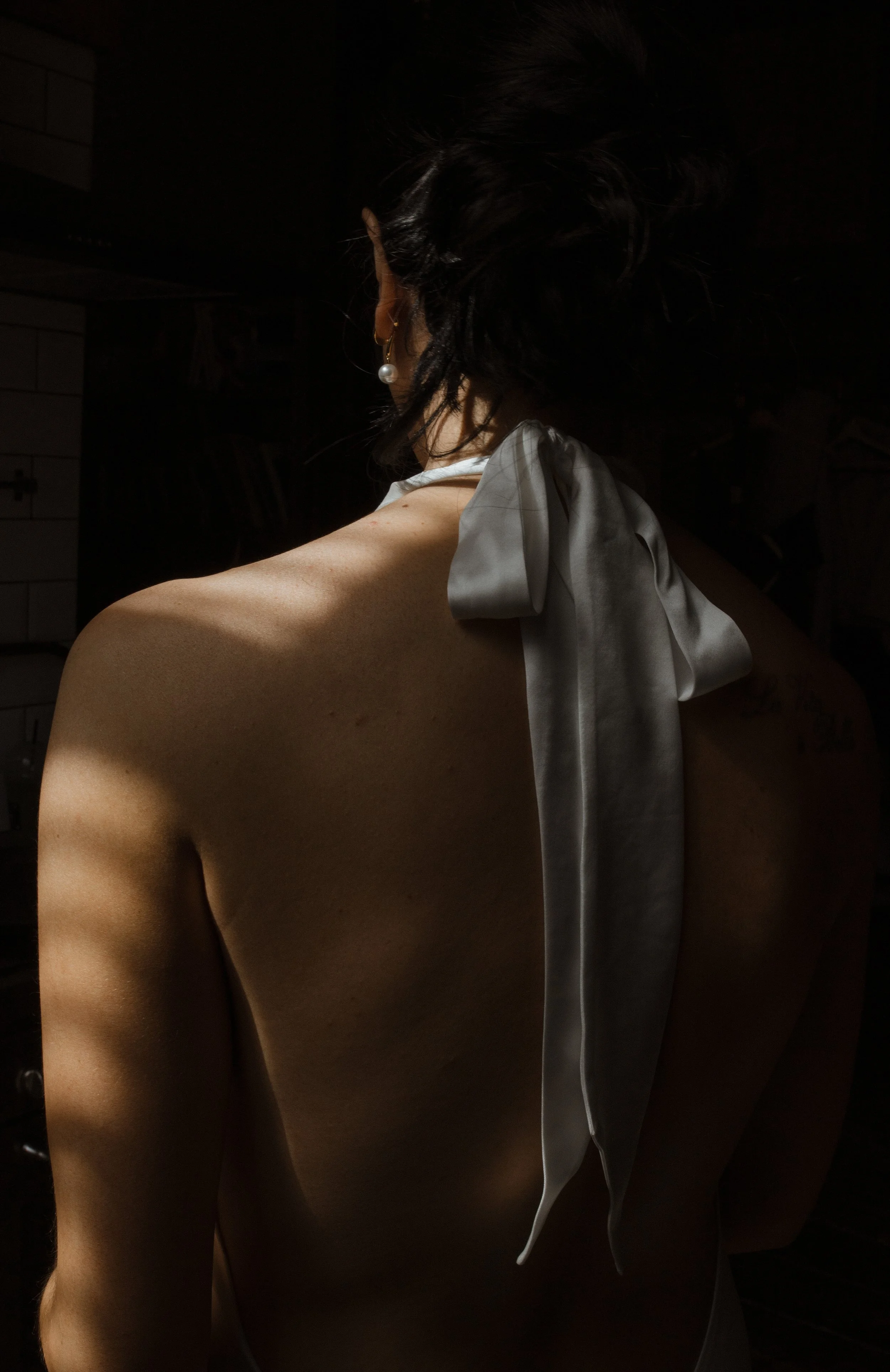 Back of a woman with dark hair wearing pearl earrings and a white satin ribbon tied in a bow, in a dimly lit room.