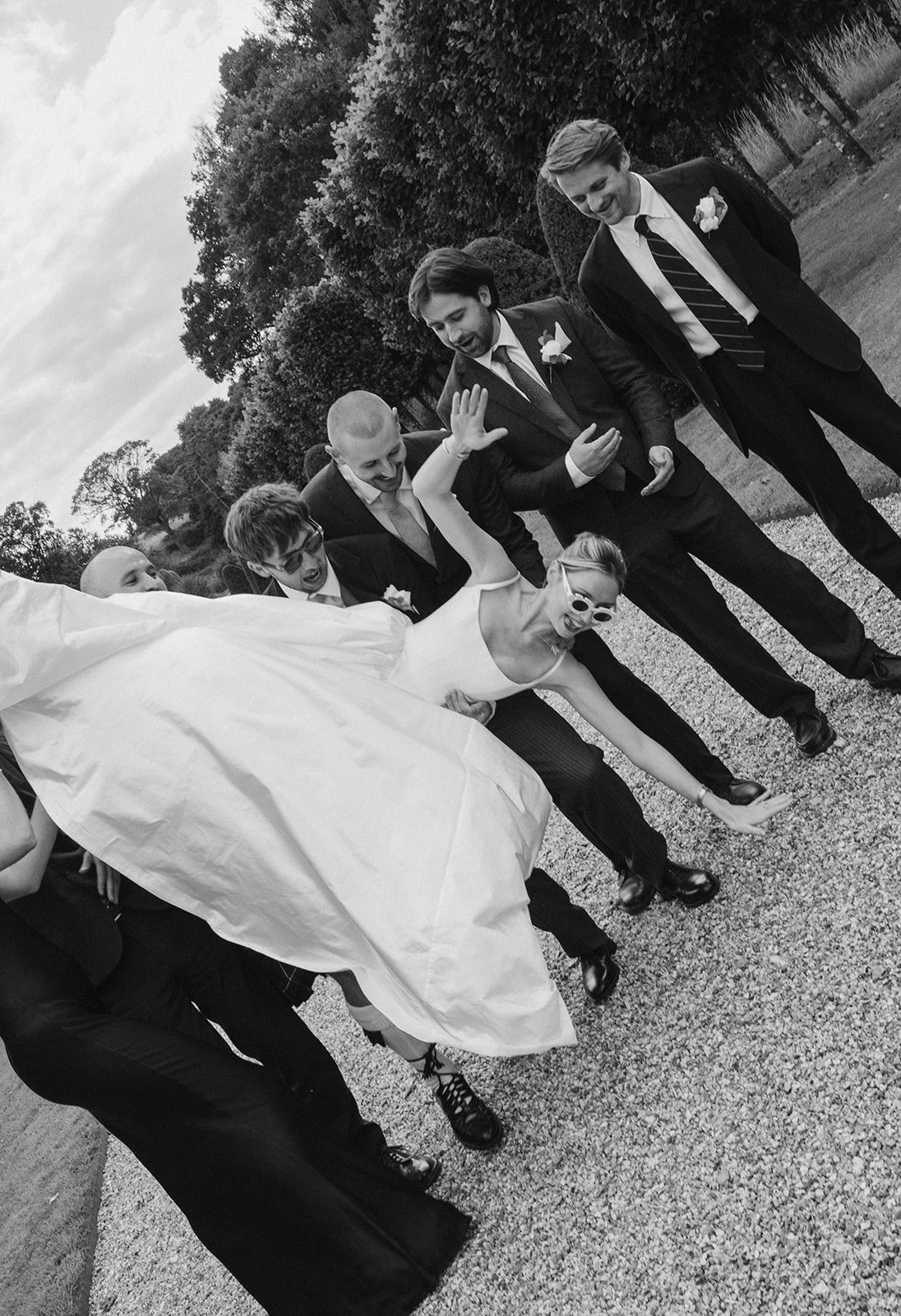A black and white photo of a bride and her wedding party, with the bride in a white dress and sunglasses, being playful and leaning forward while surrounded by men in suits. The setting is outdoors with trees and a gravel path.