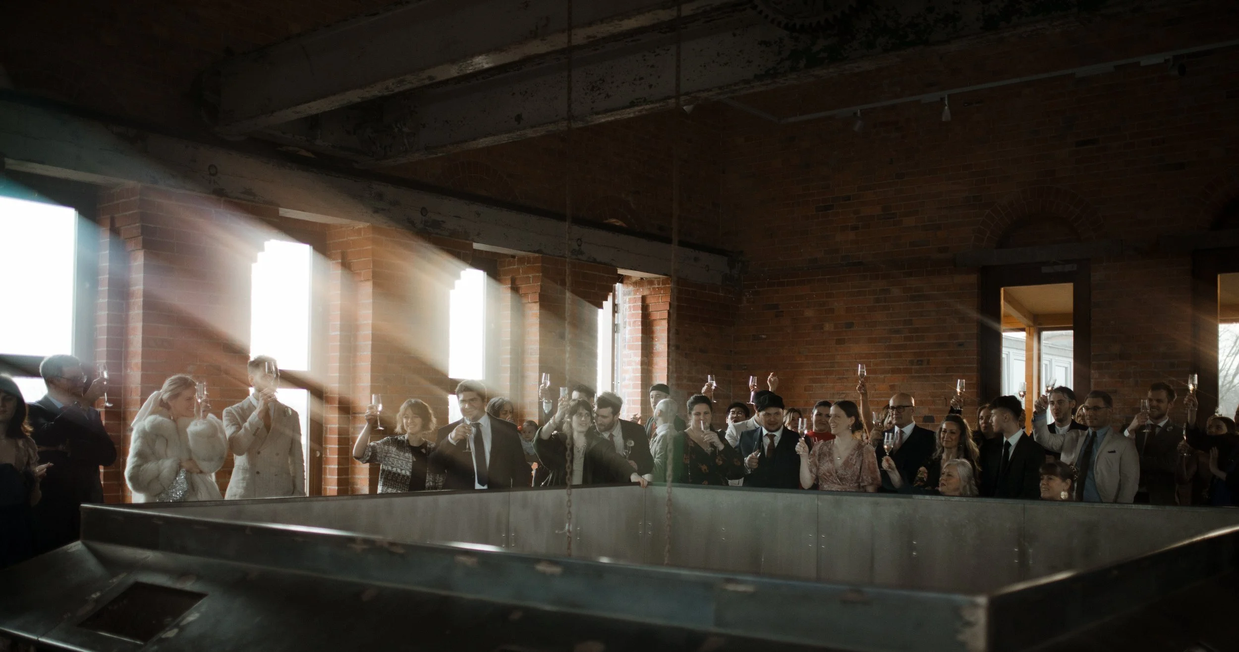 Group of people at a celebration or toast inside a brick-walled venue, some holding glasses raised in a toast, with sunlight streaming through large windows.
