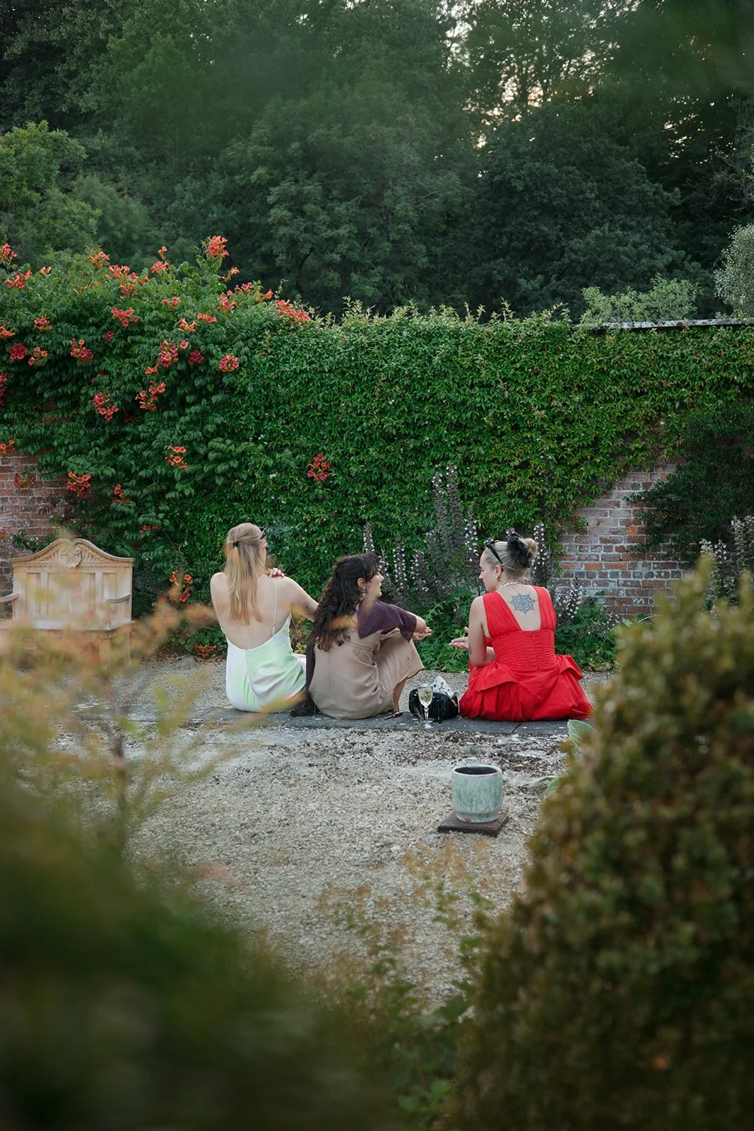 Three women sitting on the ground in a garden, engaged in conversation. The garden has lush green bushes and a brick wall in the background.