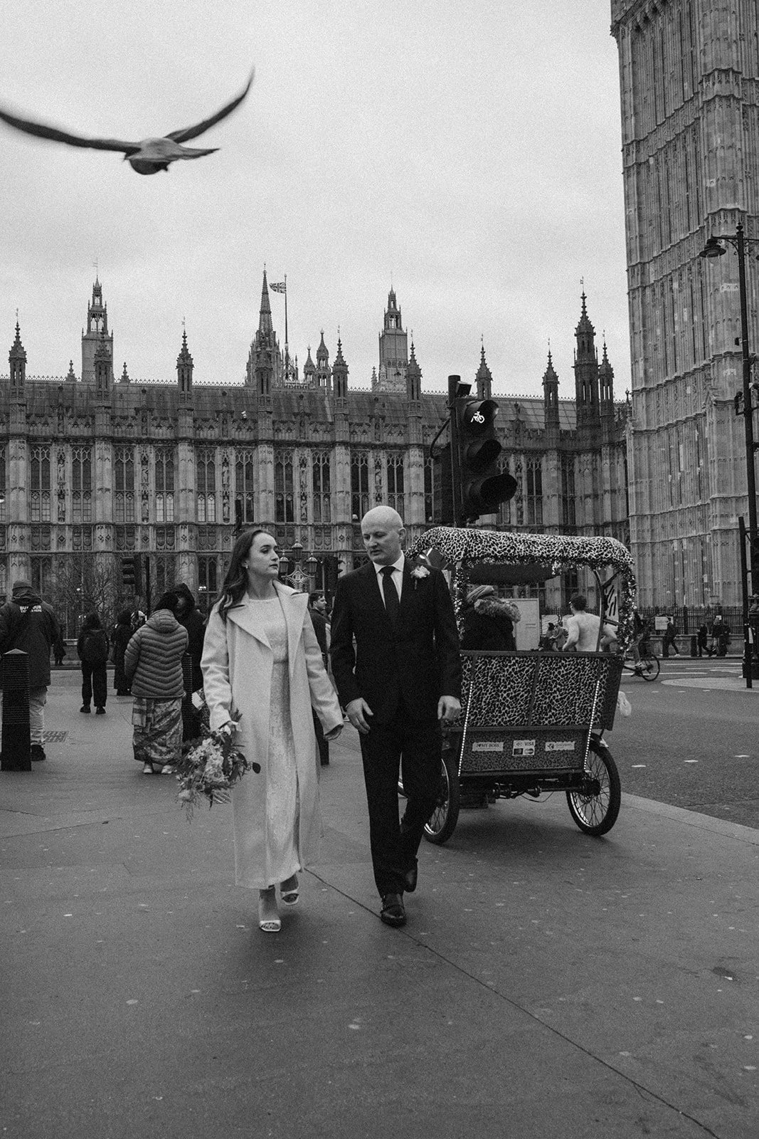 A black-and-white photo of a couple in wedding attire walking on a city street near Big Ben in London. The bride is holding a bouquet, and there are bystanders and a bicycle with a decorative carriage in the background.