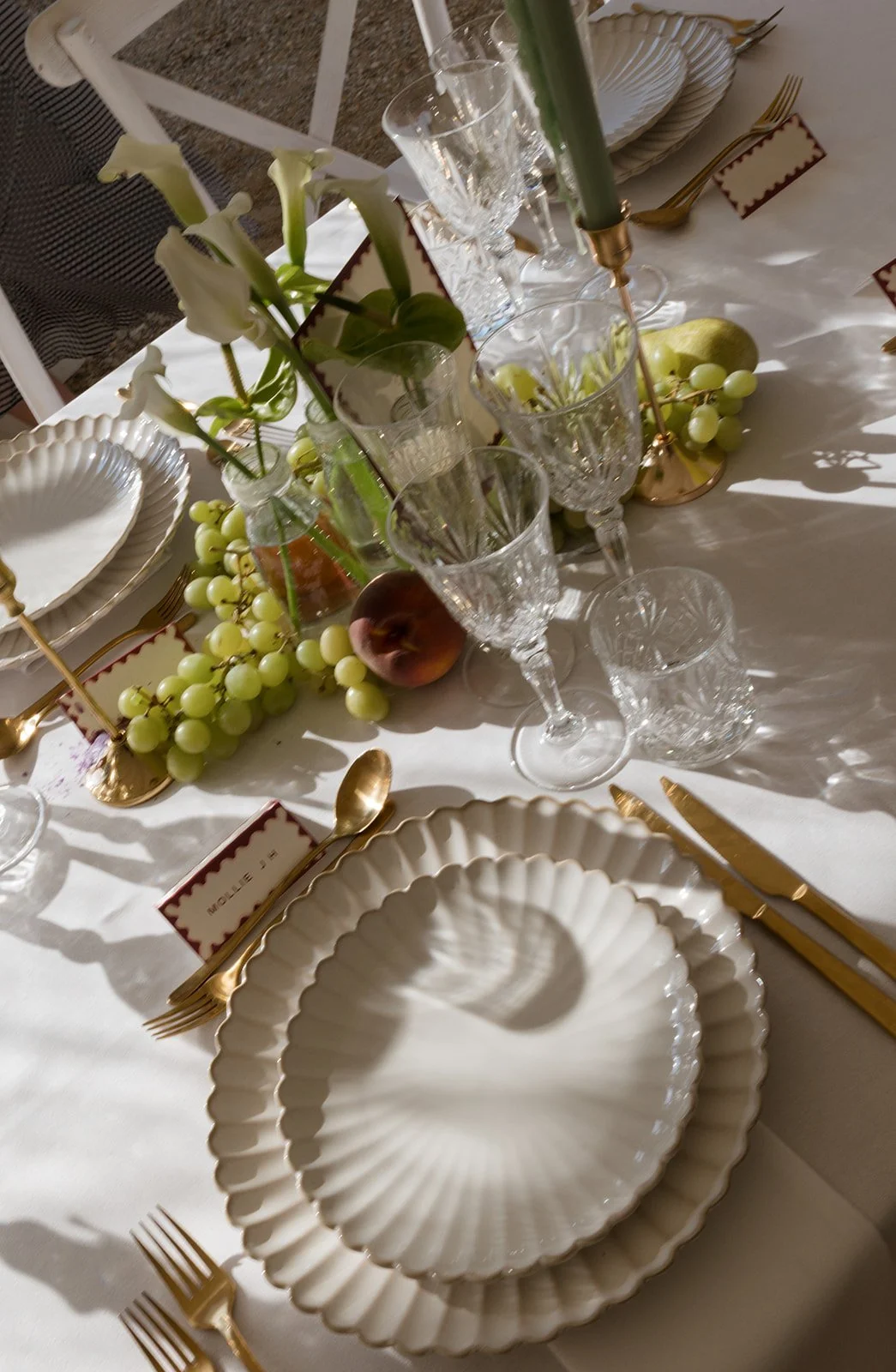 Elegant table setting with white scalloped dishes, gold utensils, crystal glasses, white floral centerpiece, and decorative fruit including grapes, pears, and a peach.