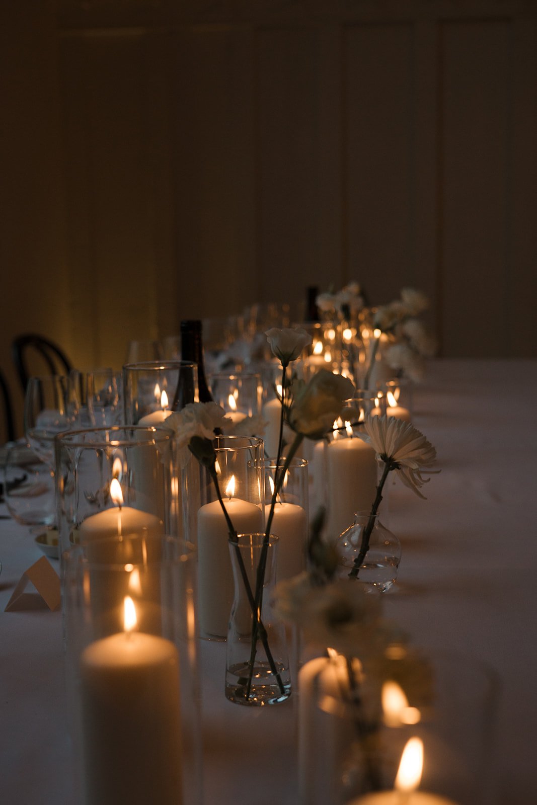 A dimly lit dining table decorated with candles in glass holders and white flowers in small vases.
