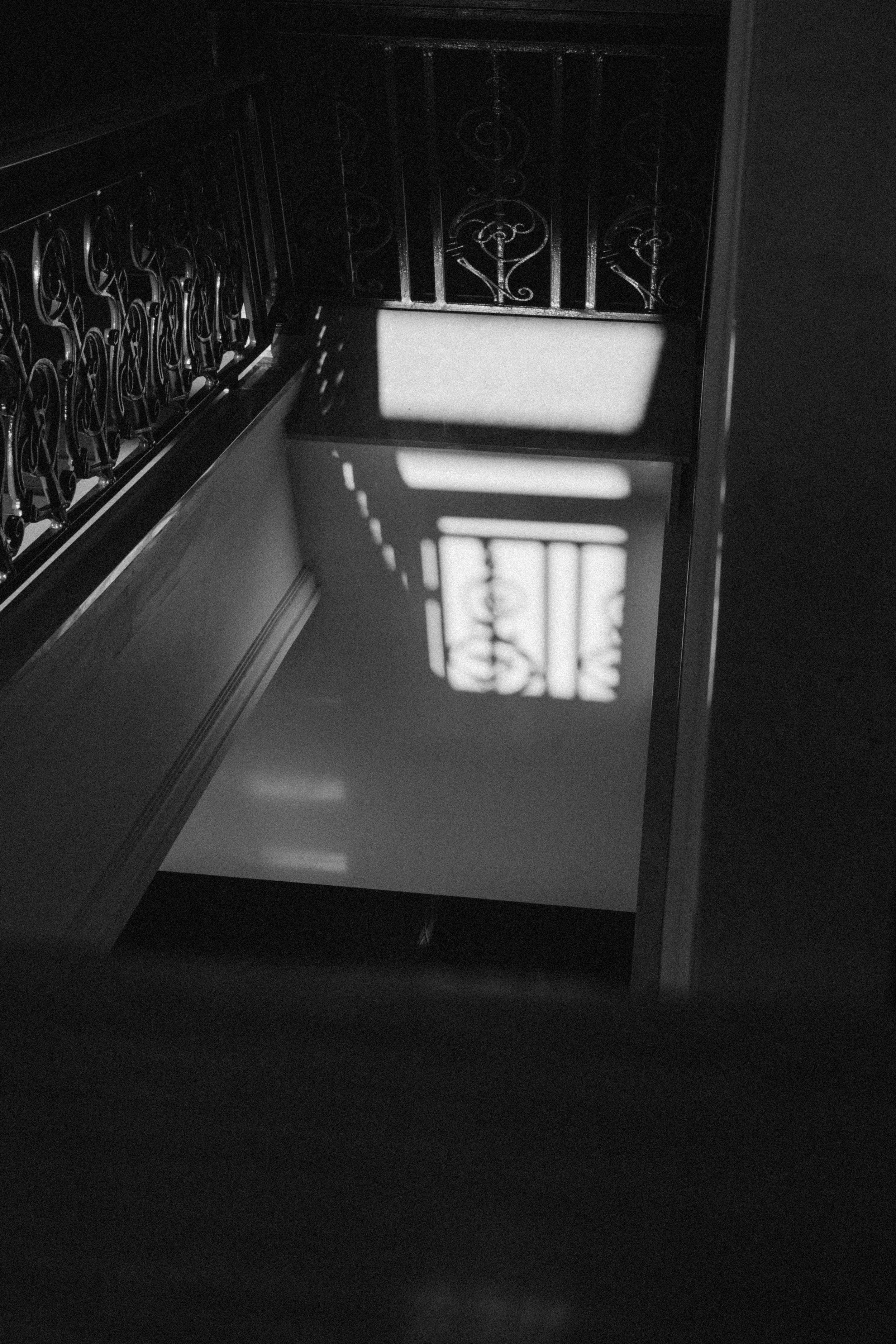 Black and white photo of a staircase with ornate railing, sunlight casting patterned shadows on the wall.