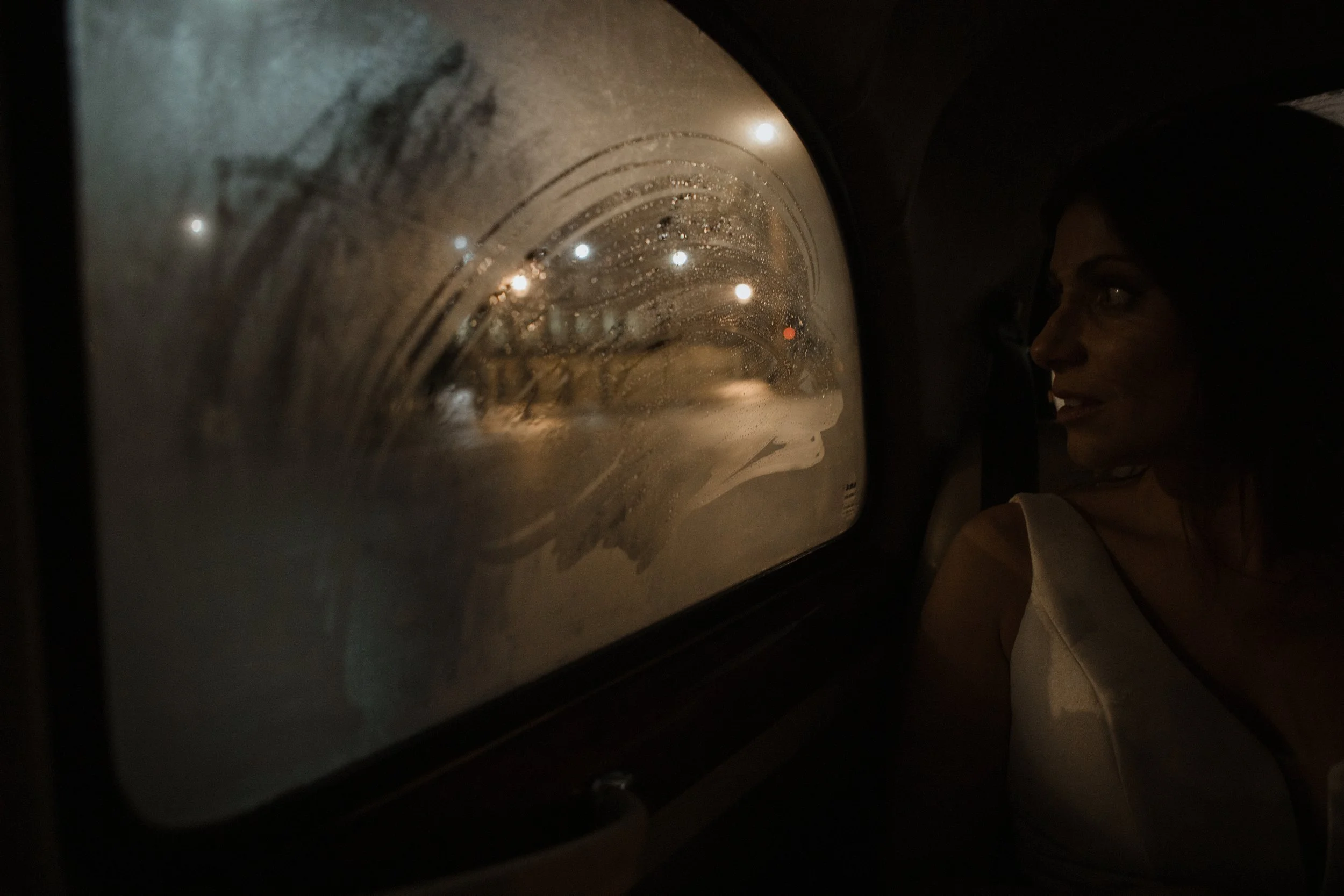 A woman sitting in a vehicle at night, looking out the window which is fogged up with streaks of moisture, illuminated by faint city lights outside.