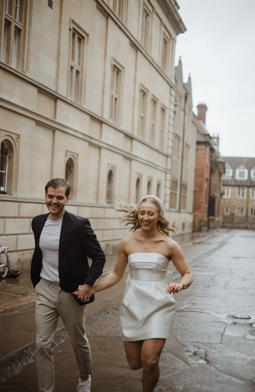 A couple, dressed in wedding attire, running hand in hand outdoors on a rainy day, with historic buildings in the background.