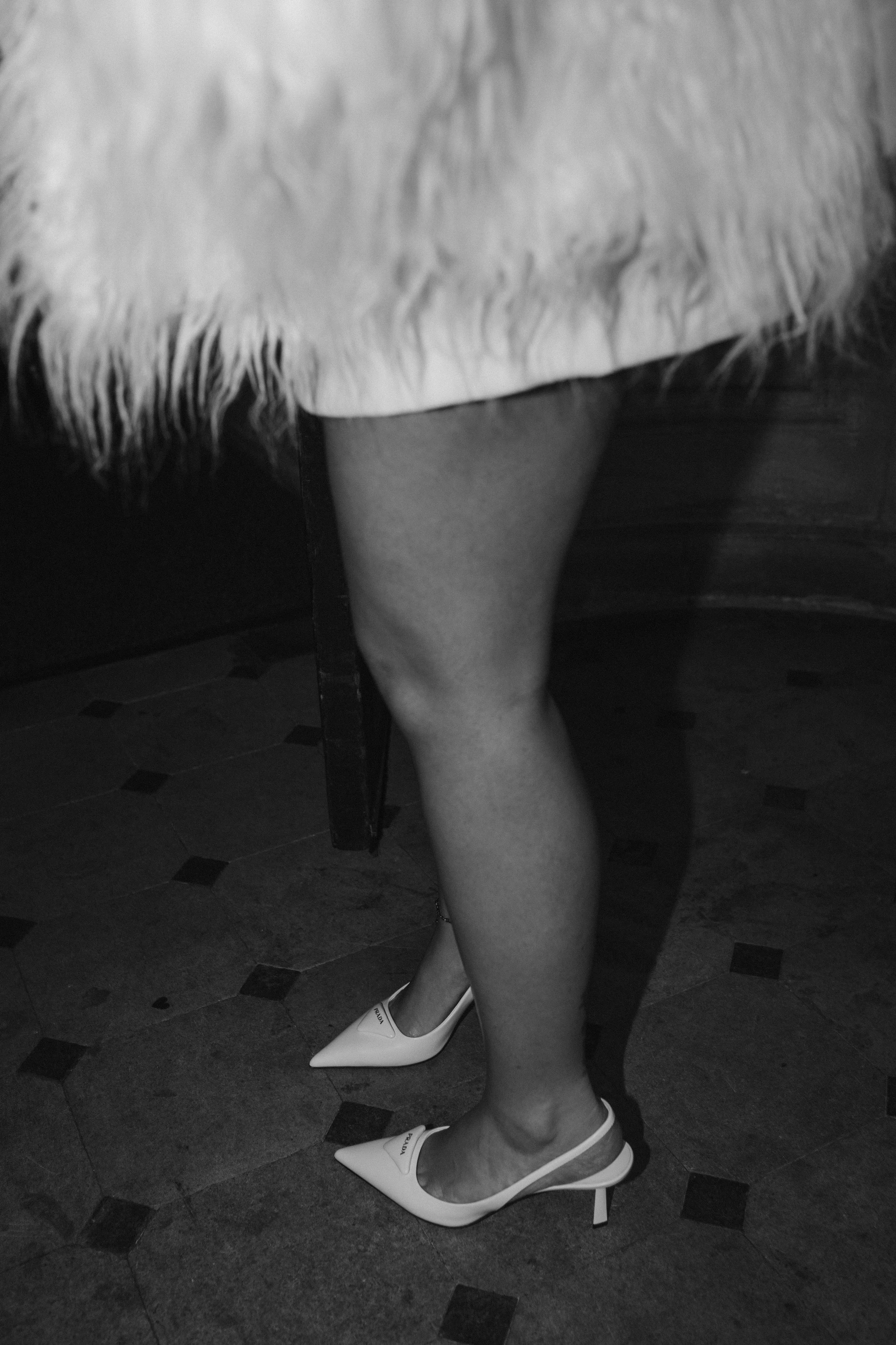 Close-up of a woman's legs in high heels and a fur coat, with part of a chair and a tiled floor visible.