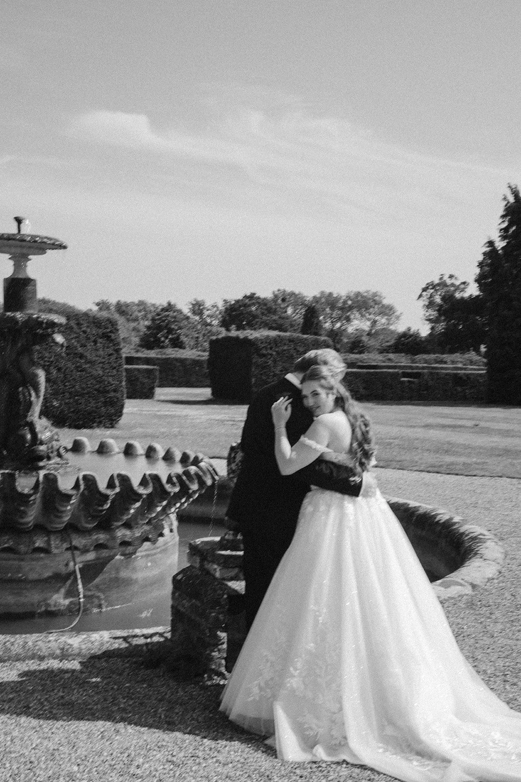 A bride and groom embracing and smiling near a fountain outdoors, with trees and manicured bushes in the background.