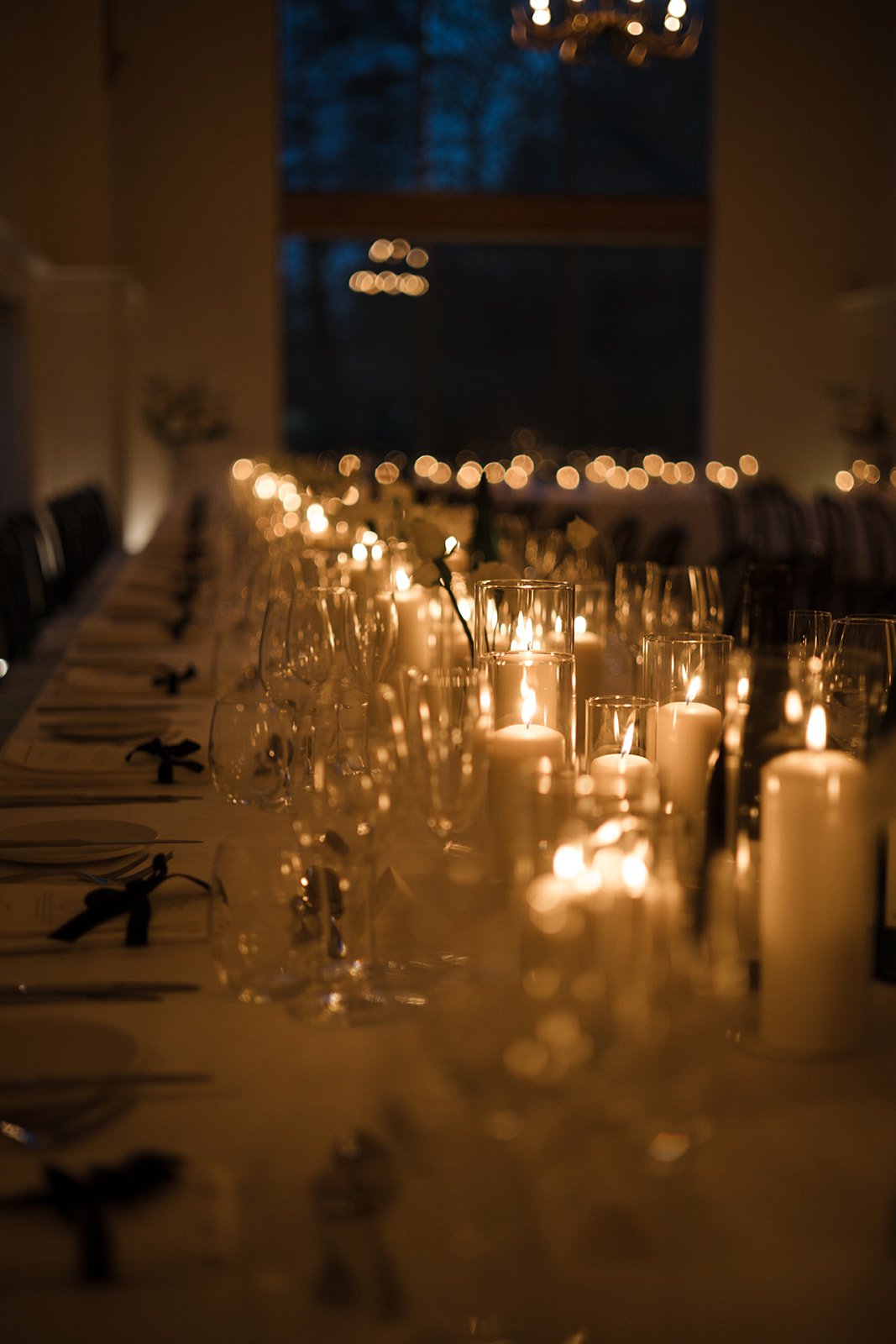 A long banquet table decorated with multiple candles in glass holders and wine glasses, set for an evening event in a dimly lit room.
