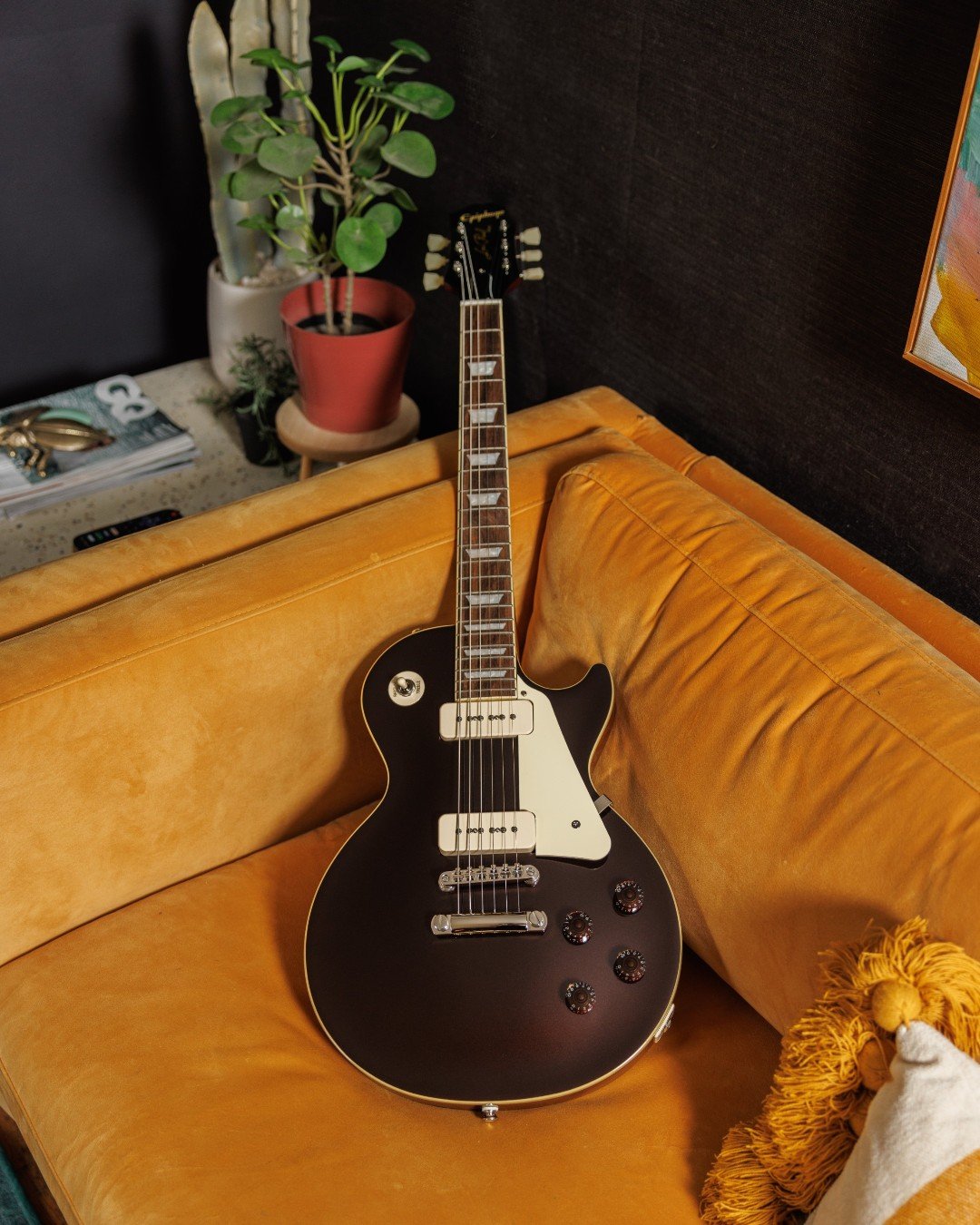 Electric guitar on a mustard yellow couch surrounded by small potted plants and decorative pillows.