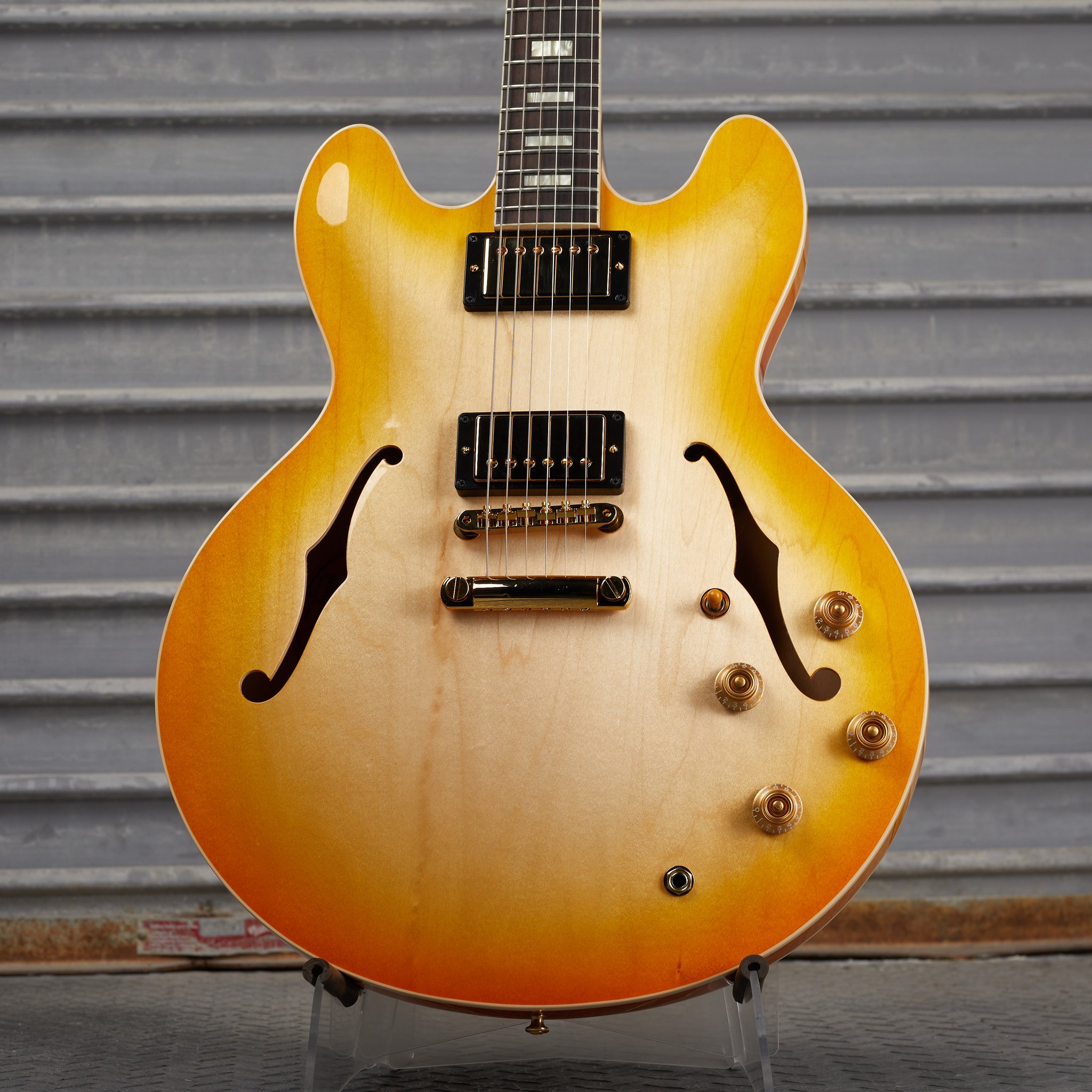 Yellow and brown semi-hollow electric guitar with two humbucker pickups and control knobs, leaning against a corrugated metal background.