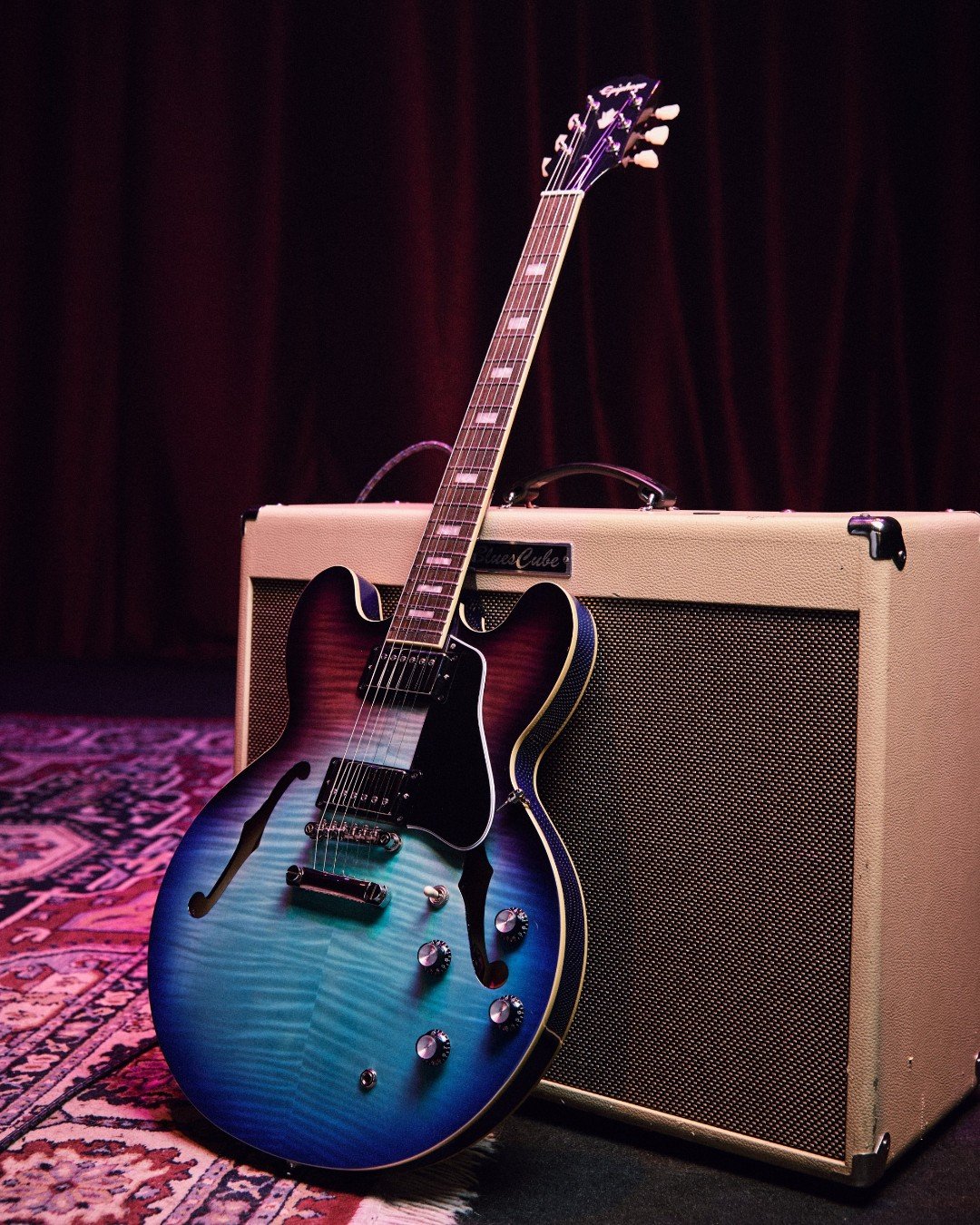 A blue and purple electric guitar leaning against a beige amplifier on a patterned rug with a dark curtain in the background.