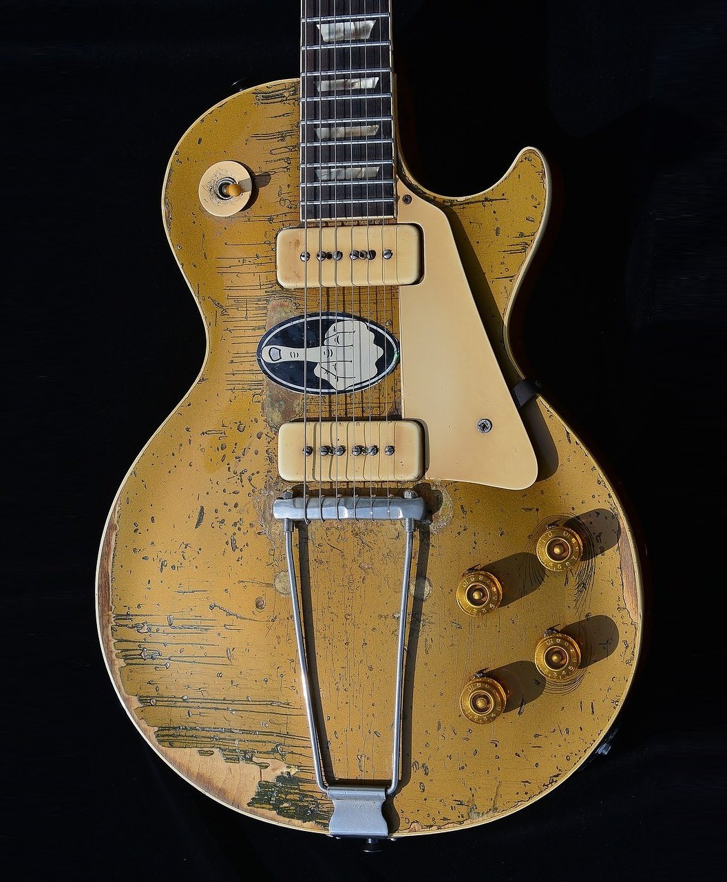 Vintage electric guitar with gold top and worn finish, featuring P-90 pickups, a trapeze tailpiece, and control knobs on a dark background.