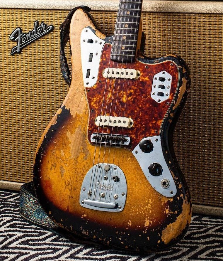 A vintage electric guitar with a sunburst finish and worn, distressed body, featuring a tortoiseshell pickguard and dual pickups, leaning against a Fender amplifier.