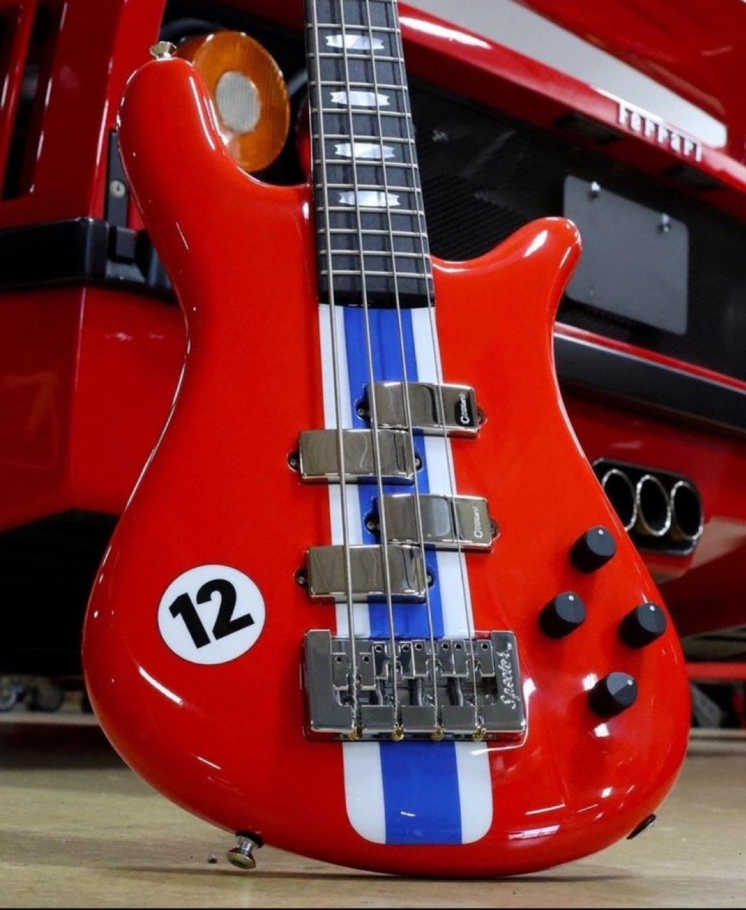 Red electric bass guitar with blue and white racing stripe, number 12 decal, in front of red sports car.