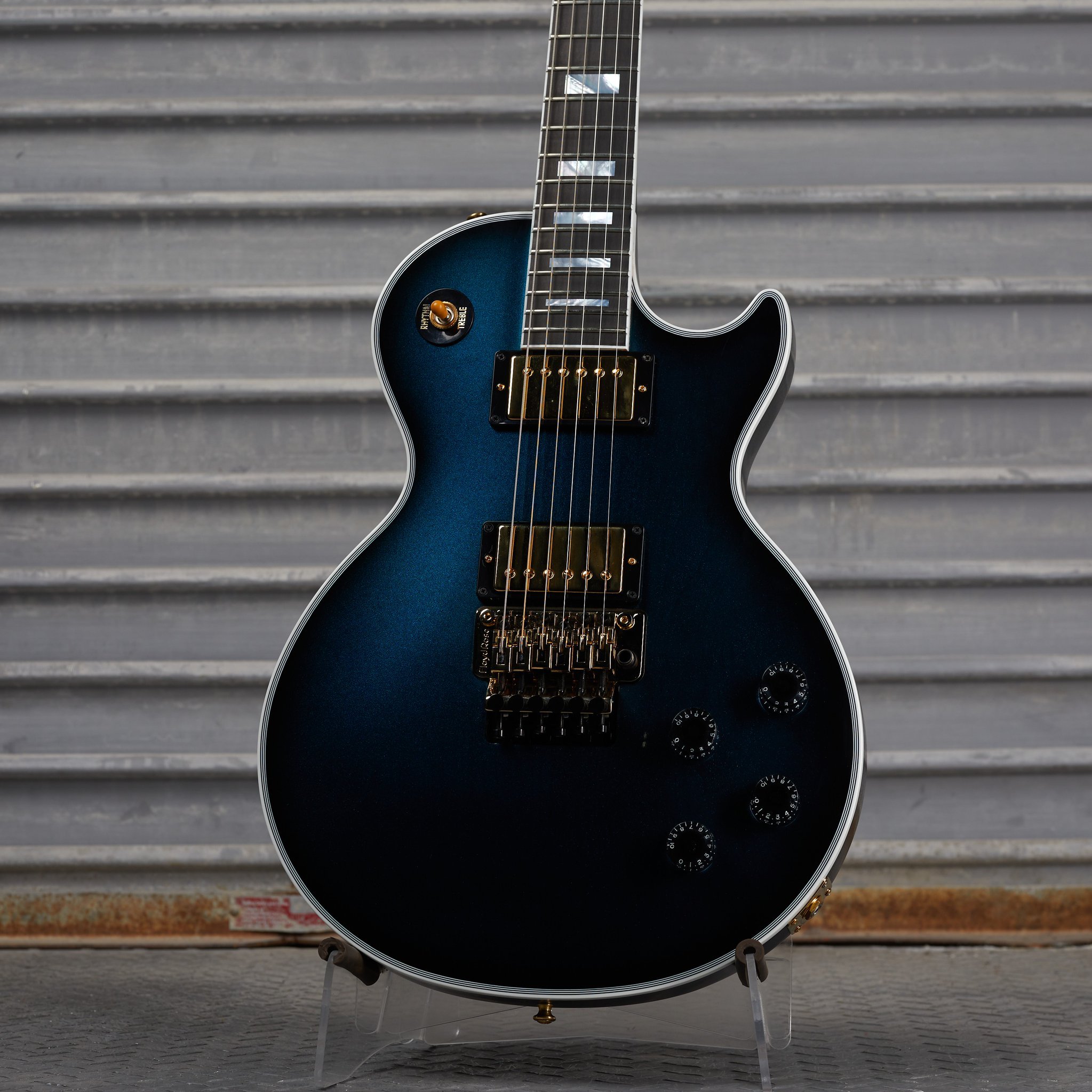 Close-up of a blue electric guitar with gold hardware and four control knobs, resting against a metal shutter background.