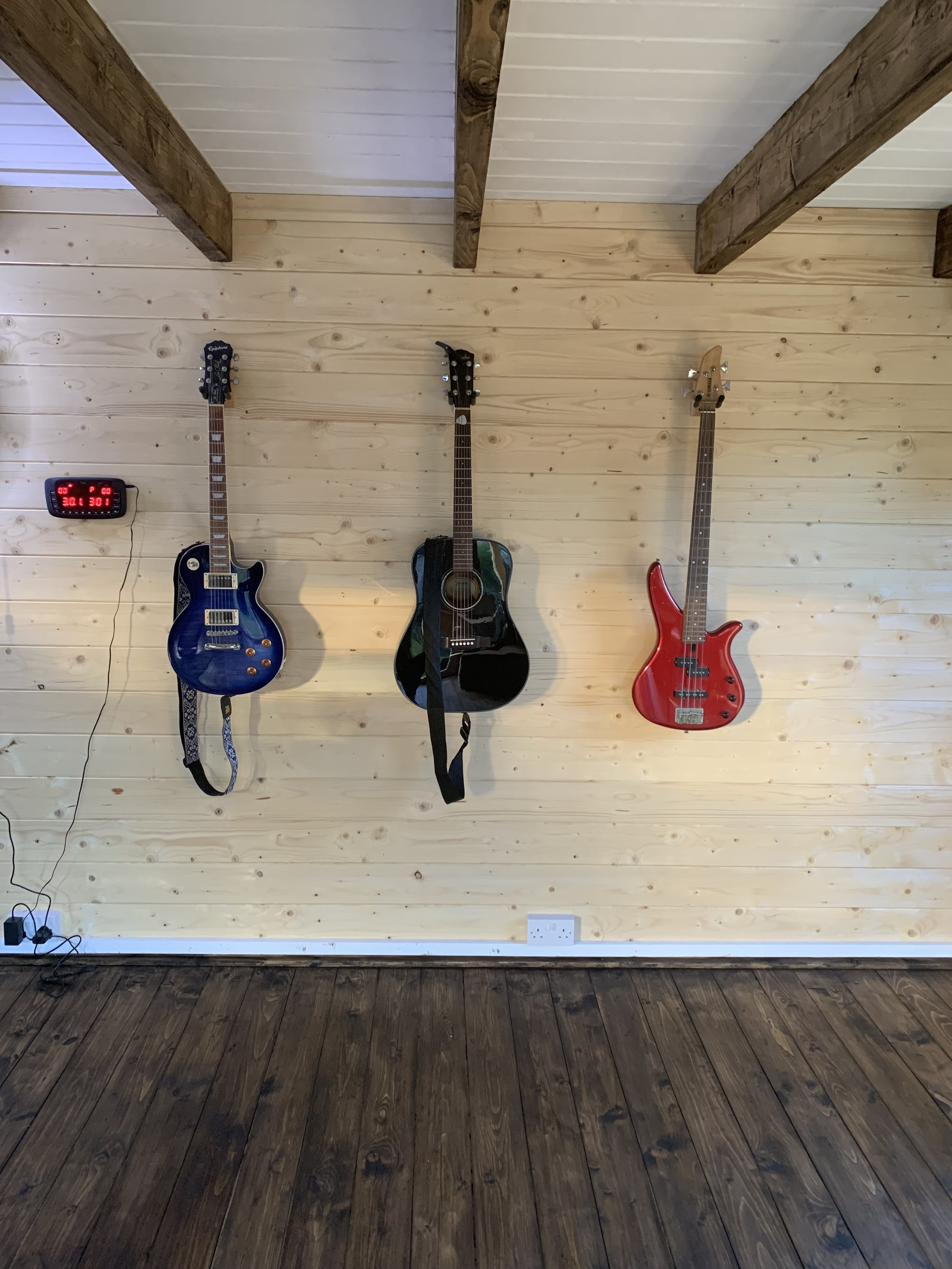 Three guitars hanging on a wooden wall in a room with a wooden floor and exposed beams. A blue electric guitar, a black acoustic guitar, and a red bass guitar are displayed. A digital clock is visible on the left side of the wall.