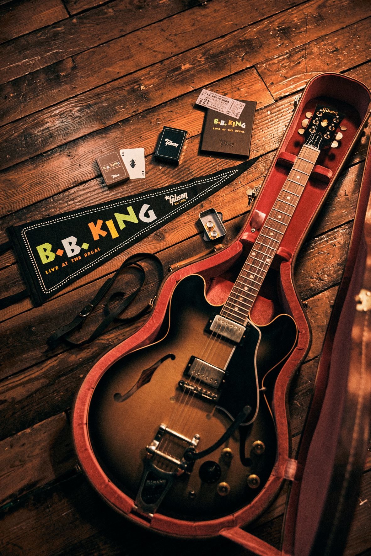 Electric guitar in a red-lined case on a wooden floor, surrounded by B.B. King memorabilia like a pennant, playing cards, picks, and concert items.