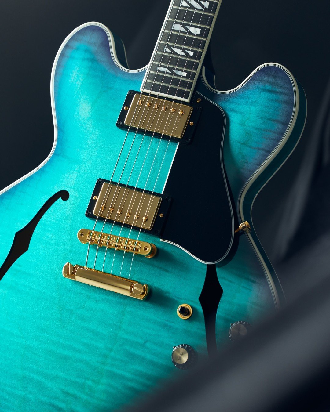 Close-up of a turquoise semi-hollow electric guitar with gold hardware, black pickguard, and visible f-holes.
