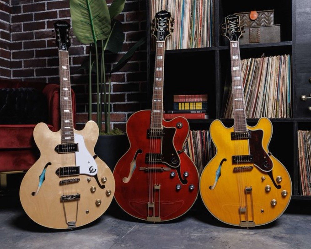 Three Epiphone semi-hollow electric guitars in natural, red, and yellow finishes, displayed in a room with a brick wall, vinyl records, and a potted plant.