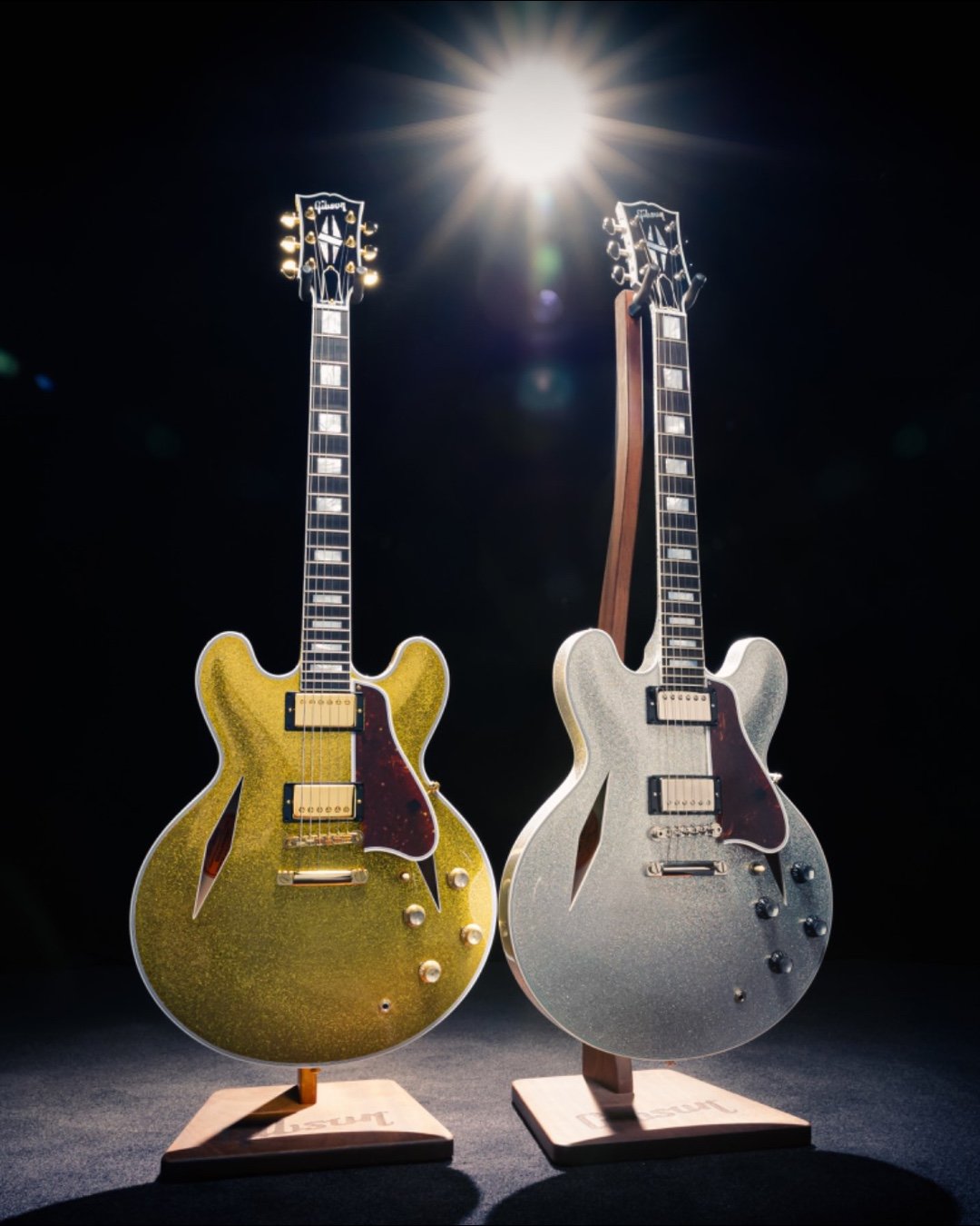 Two semi-hollow electric guitars with gold and silver sparkling finishes, displayed on stands in a dimly lit setting with a bright spotlight above.