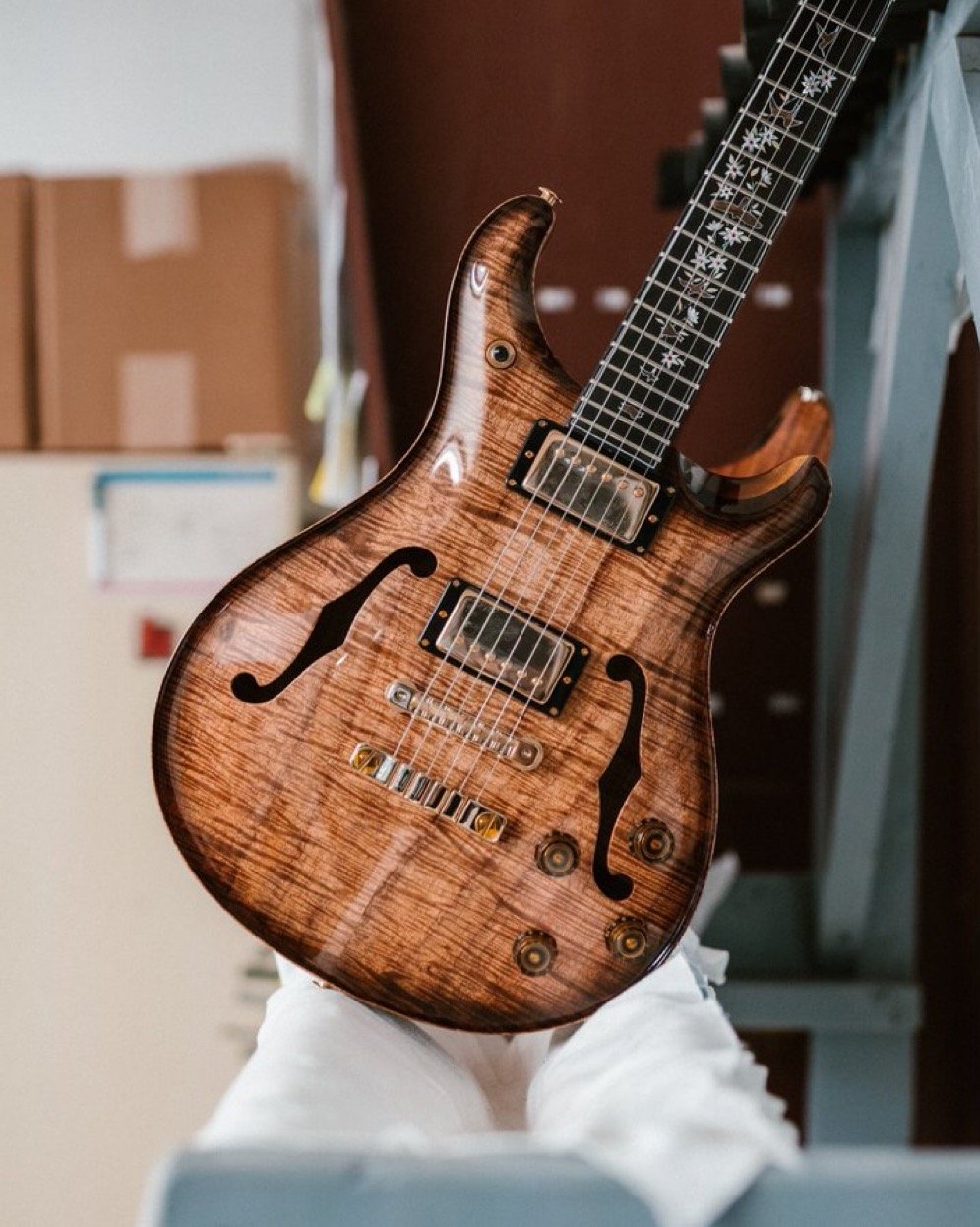 A semi-hollow electric guitar with a sunburst finish leaning against a structure, featuring a flamed maple top and intricate inlays on the fretboard.