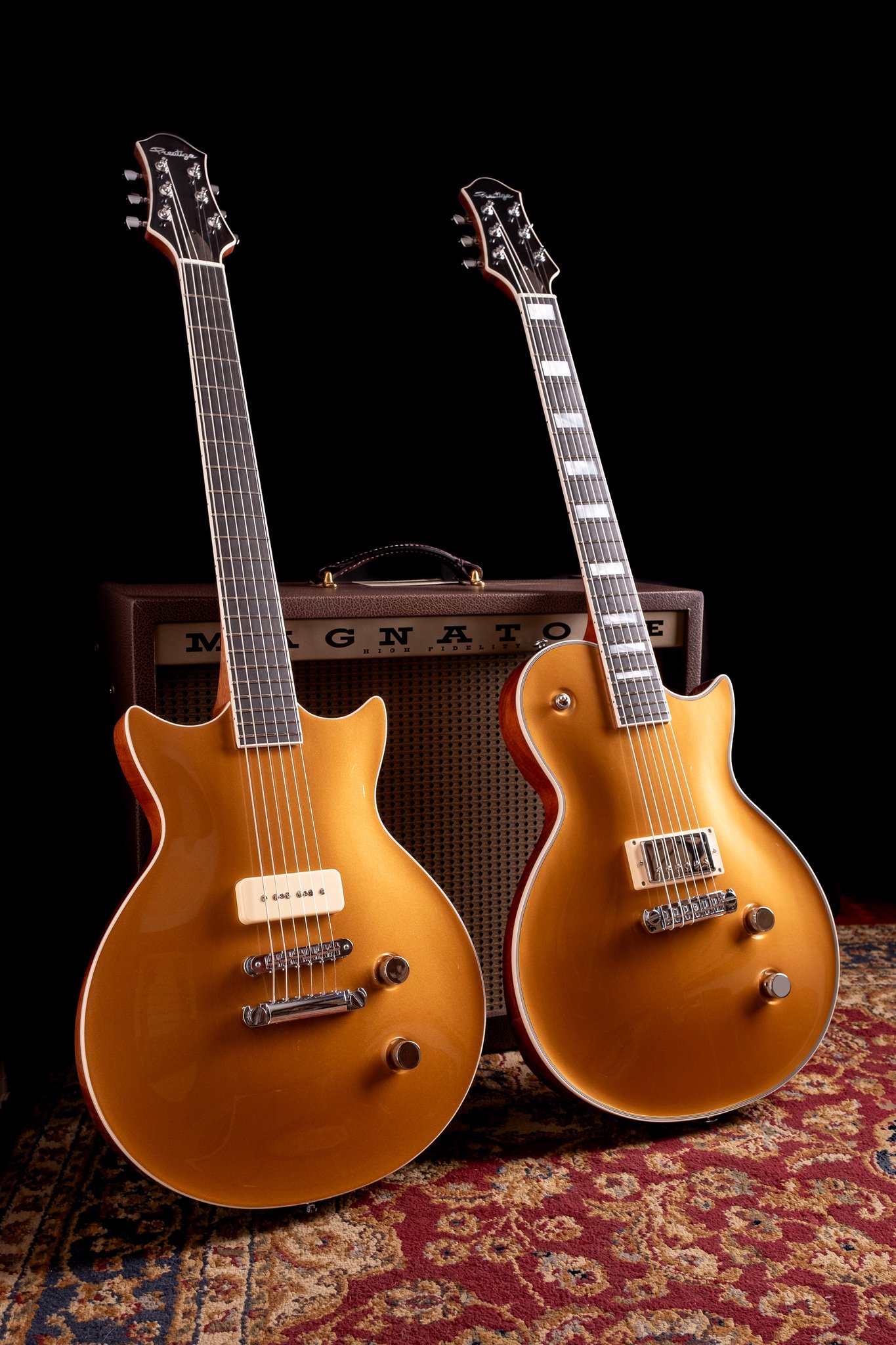 Two gold electric guitars standing in front of a vintage-style amplifier on a patterned rug.