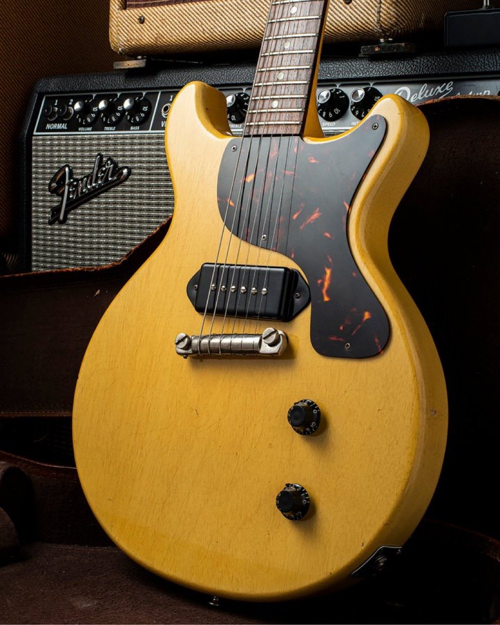 A vintage electric guitar with a yellow body and a tortoiseshell pickguard, positioned in front of a Fender amp.