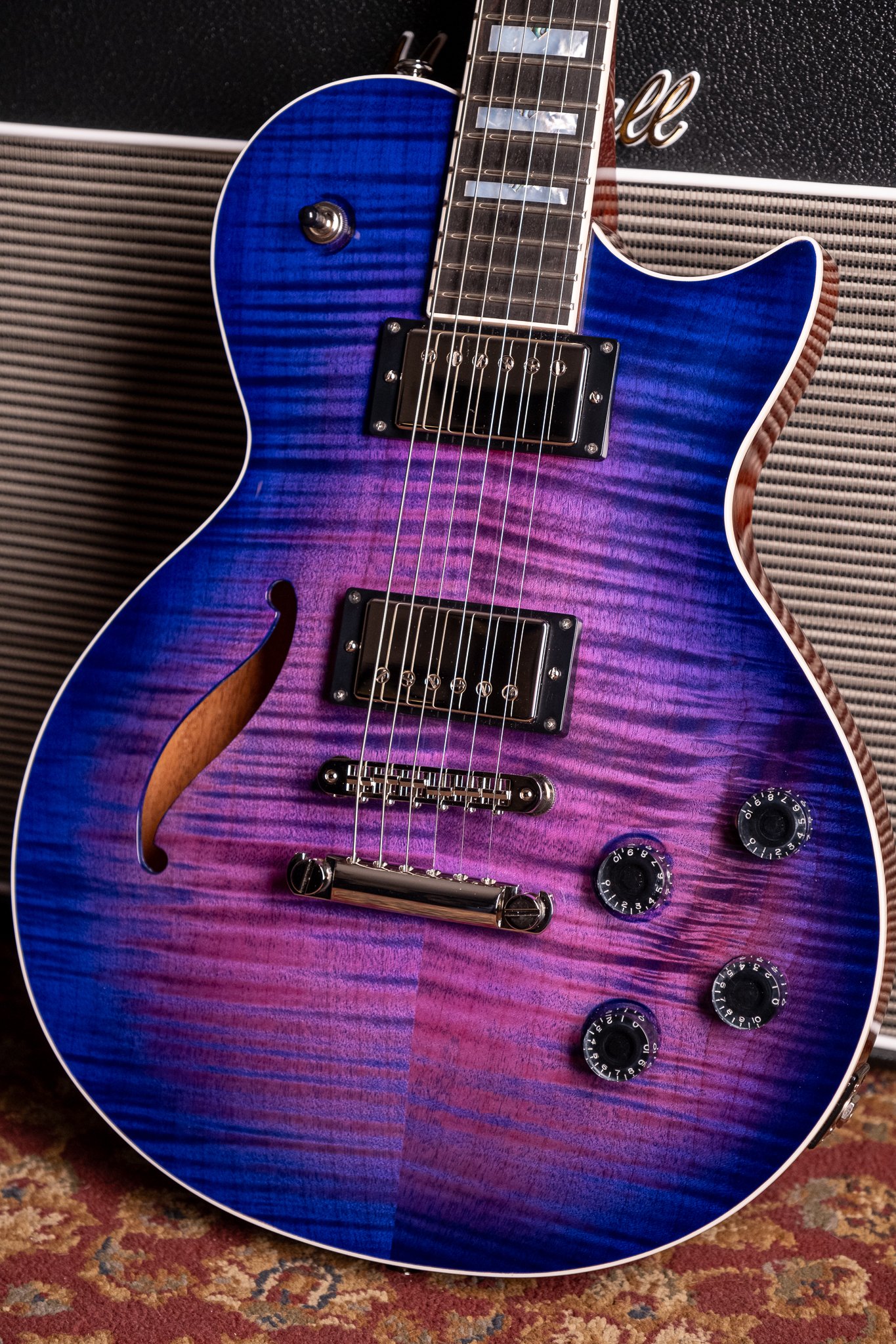 Electric guitar with blue and purple finish, featuring a semi-hollow body and humbucker pickups, against an amplifier.