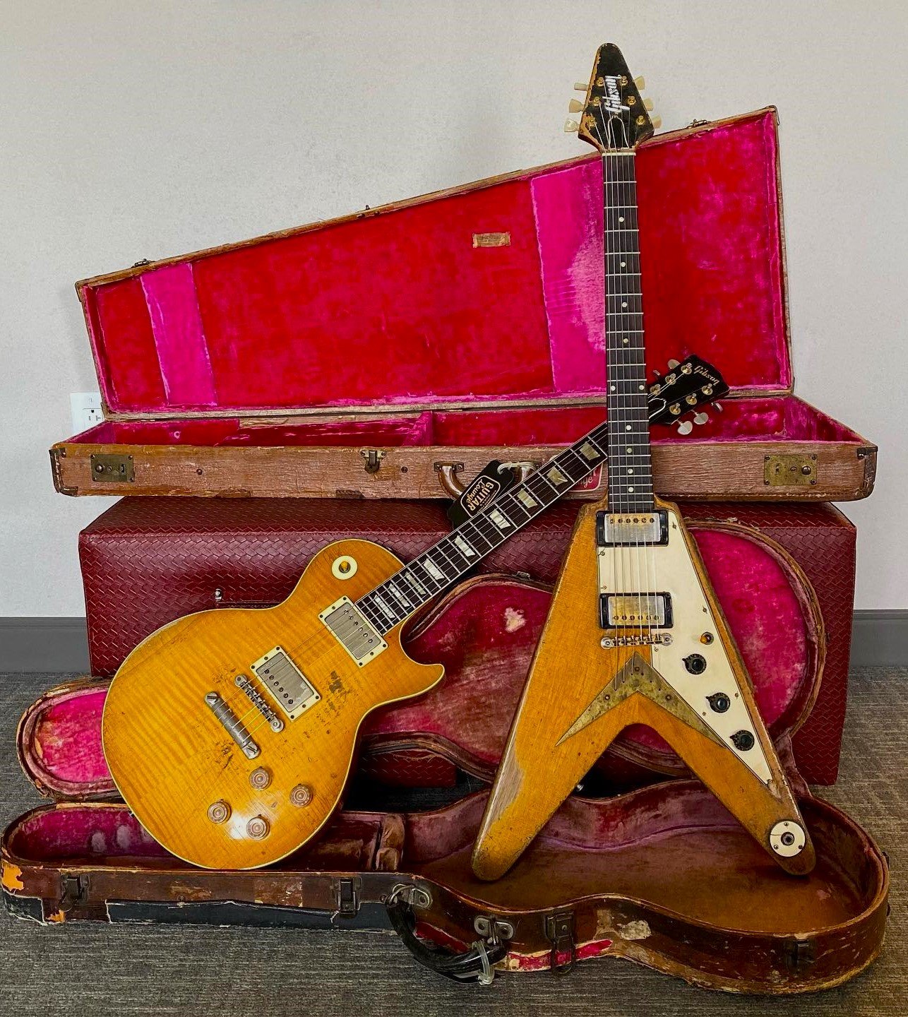 Two vintage electric guitars, a Gibson Les Paul and a Gibson Flying V, leaning against open guitar cases with red velvet lining.