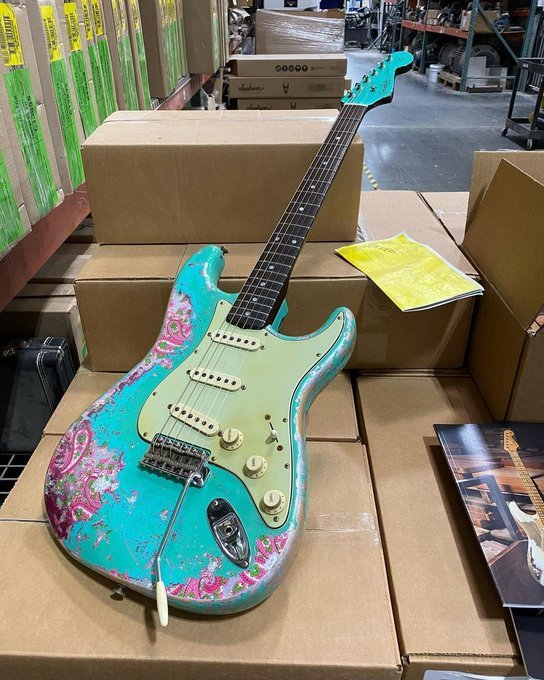 Electric guitar with turquoise and pink paisley design resting on cardboard boxes in a warehouse setting.