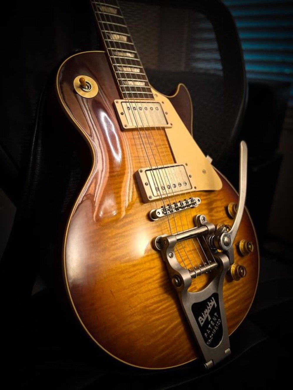 Close-up of an electric guitar with a sunburst finish and a Bigsby tremolo, set against a dark background.