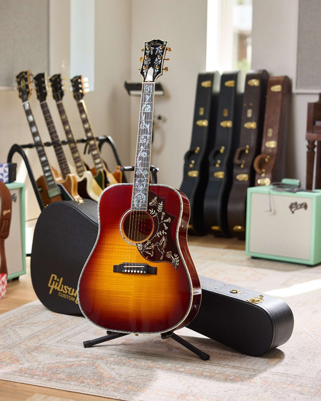 A sunburst acoustic guitar with intricate inlays and decoration on the pickguard, displayed on a stand in a room. A Gibson custom guitar case is nearby. Several electric guitars are in the background. The room has carpeted flooring and natural light.