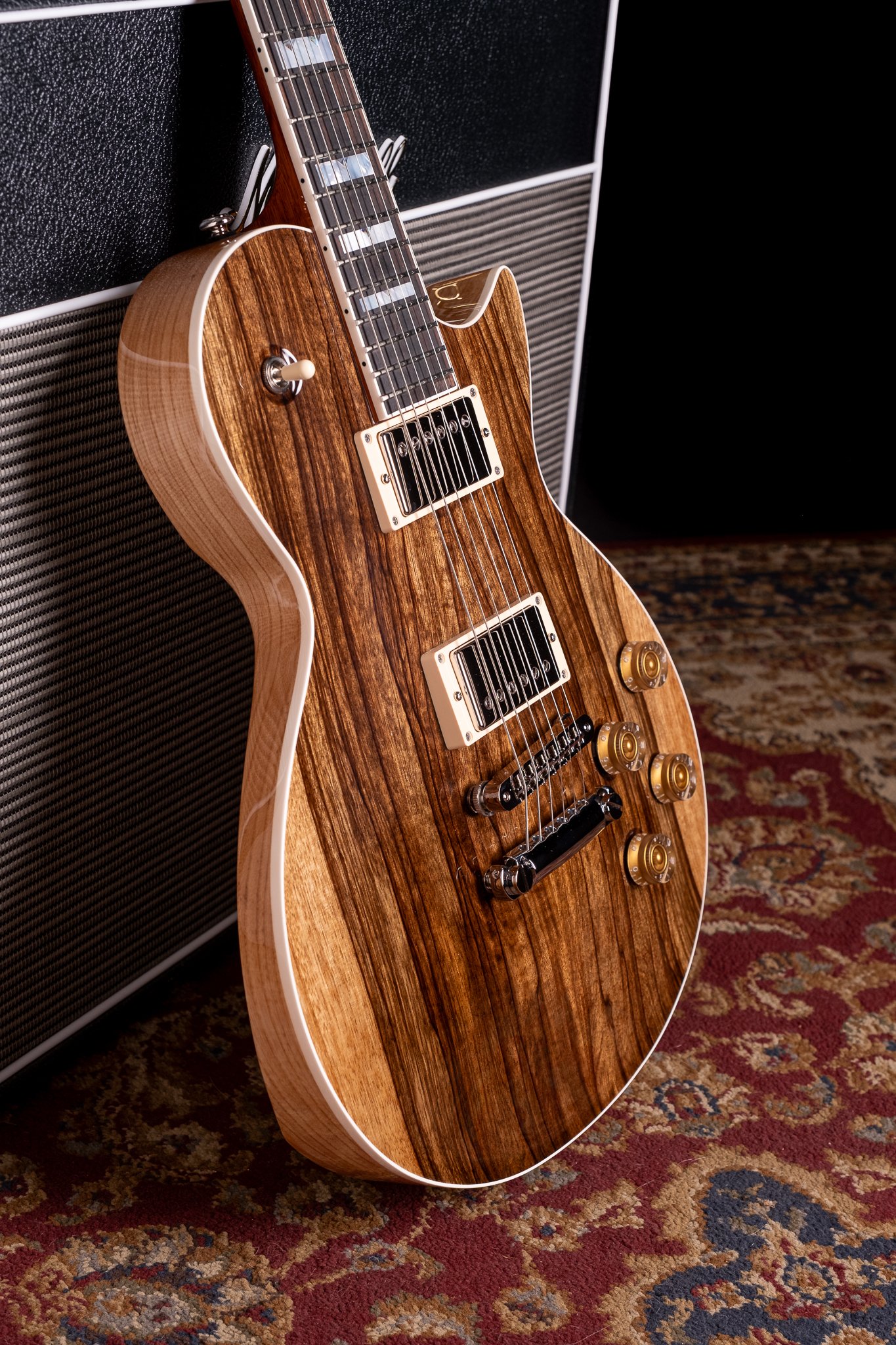 Wooden electric guitar leaning against a black amplifier on a patterned carpet.