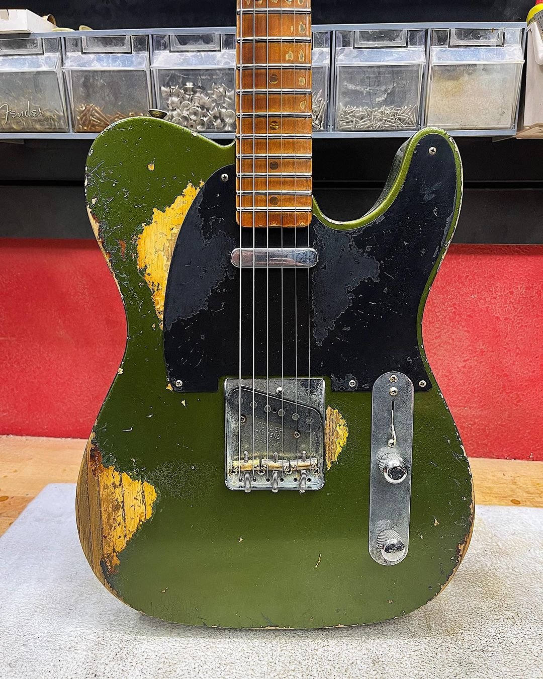A worn, vintage electric guitar with a green and yellow distressed body and black pickguard, positioned in front of storage containers filled with hardware, displayed on a red background.